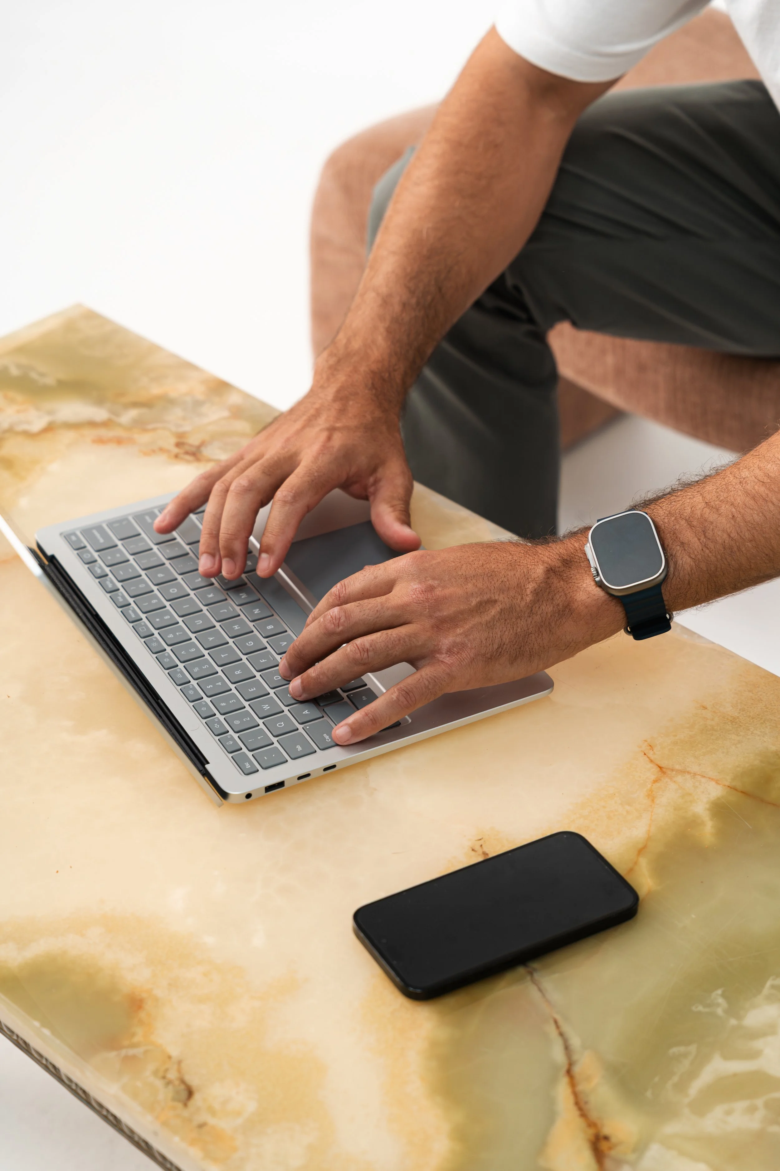 A person leaning over a laptop on a marble table, with a smartphone nearby.