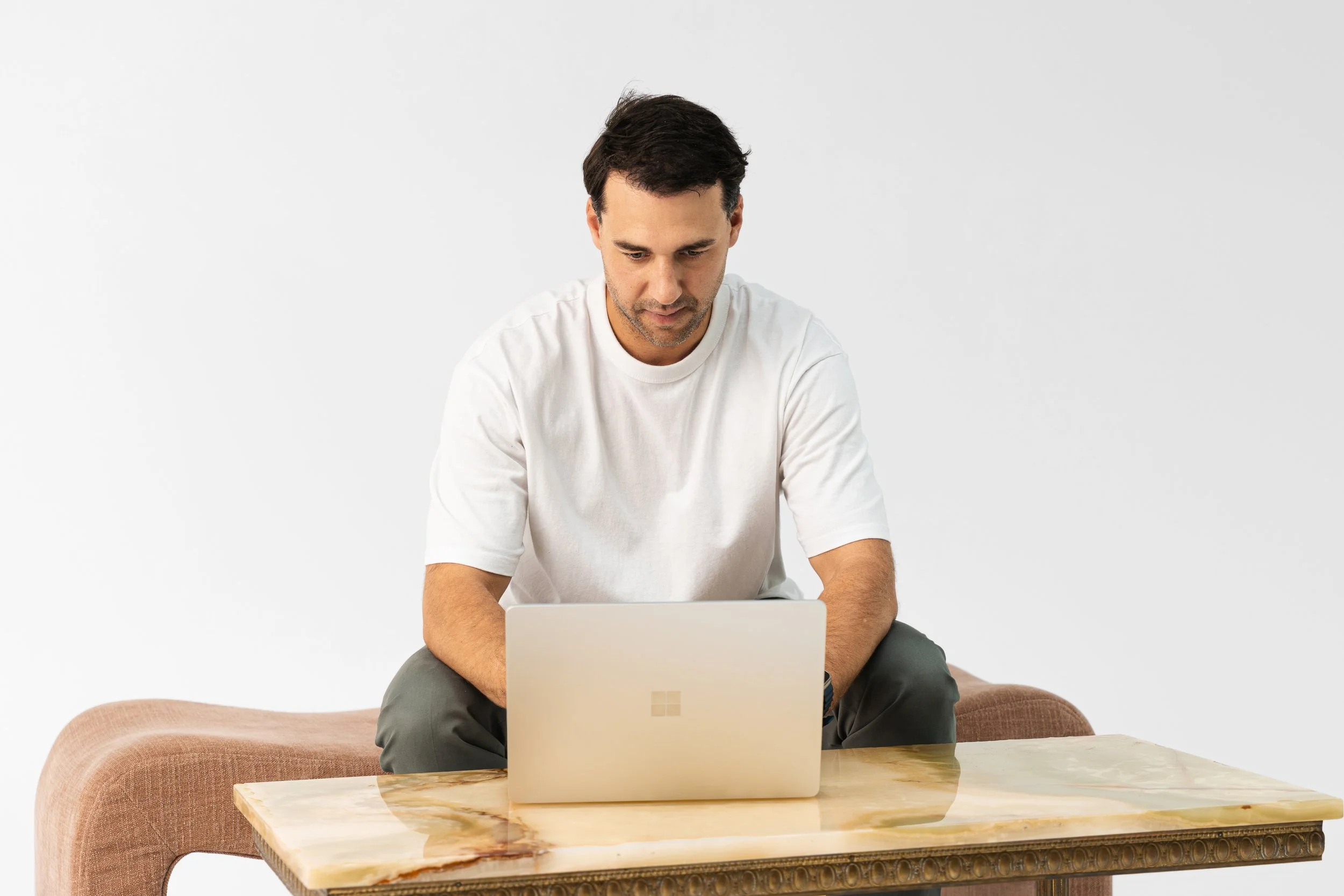 A man sitting at a wooden table working on a silver laptop with a Microsoft logo, in a minimalistic room with white walls and a beige sofa.