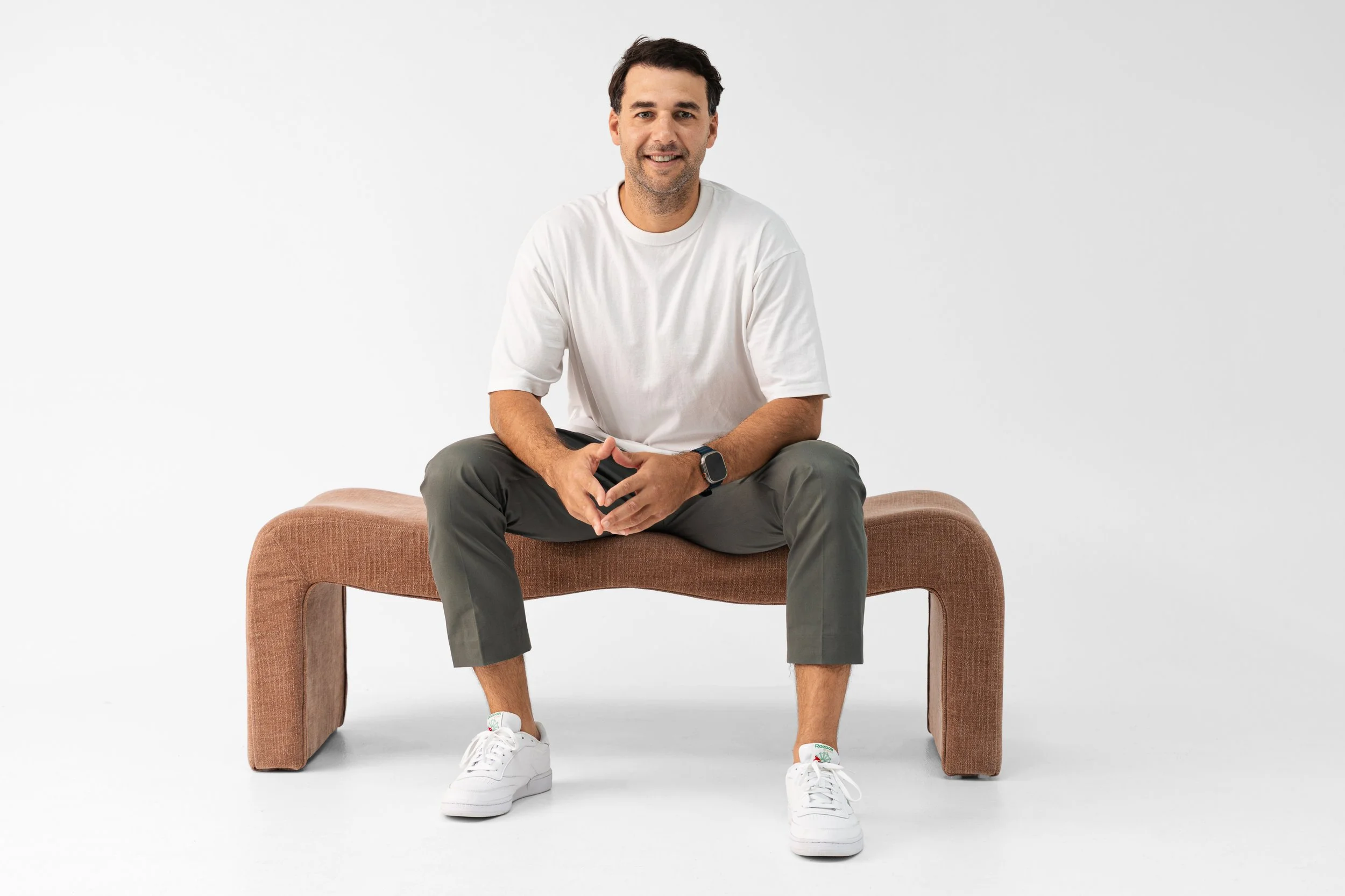 A man sitting on a tan upholstered bench against a plain white background, wearing a white t-shirt, gray pants, white sneakers, and a smartwatch, smiling at the camera.