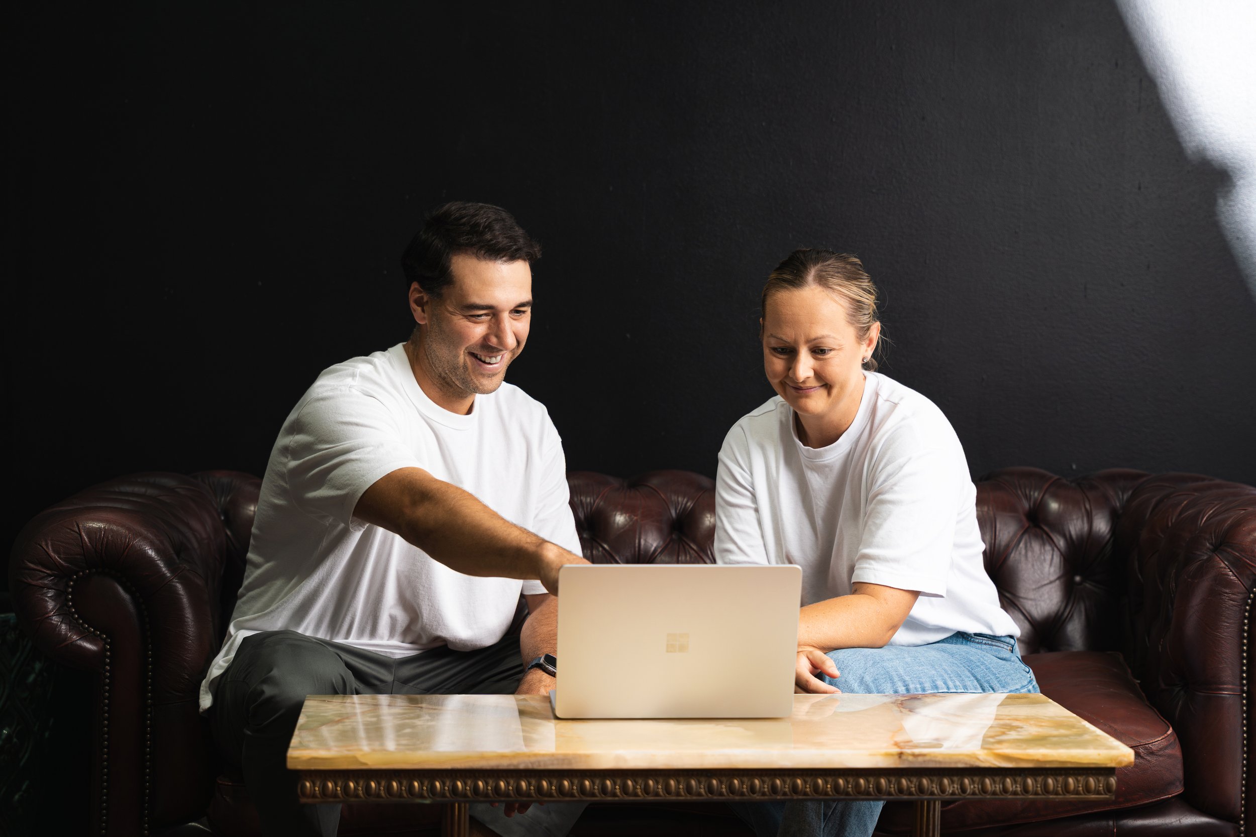 A man and woman sitting on a leather sofa, looking at a laptop on a wooden coffee table, with a dark wall background.