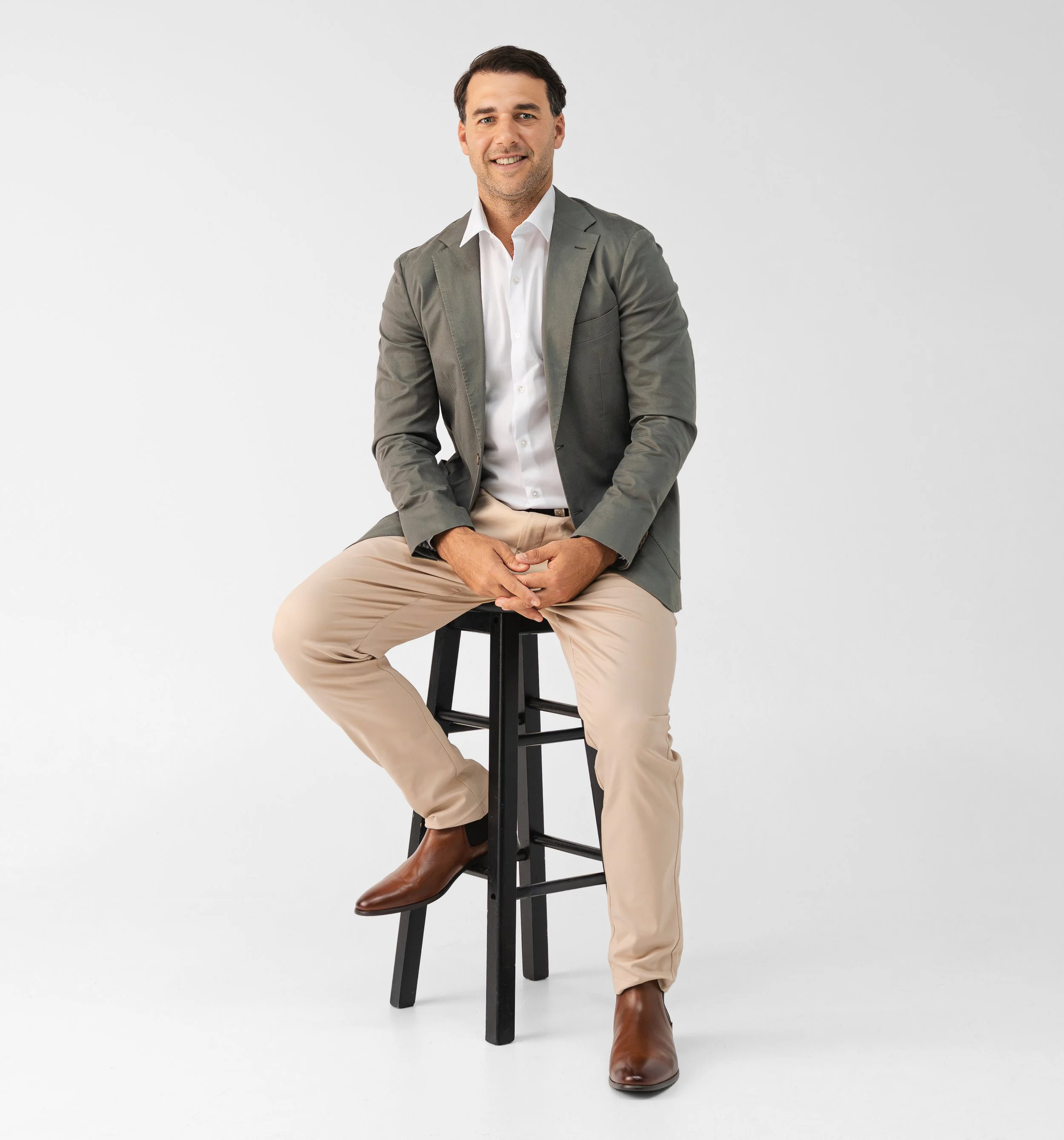 A man sitting on a black stool, dressed in a gray blazer, white shirt, beige pants, and brown boots, posed against a plain white background.