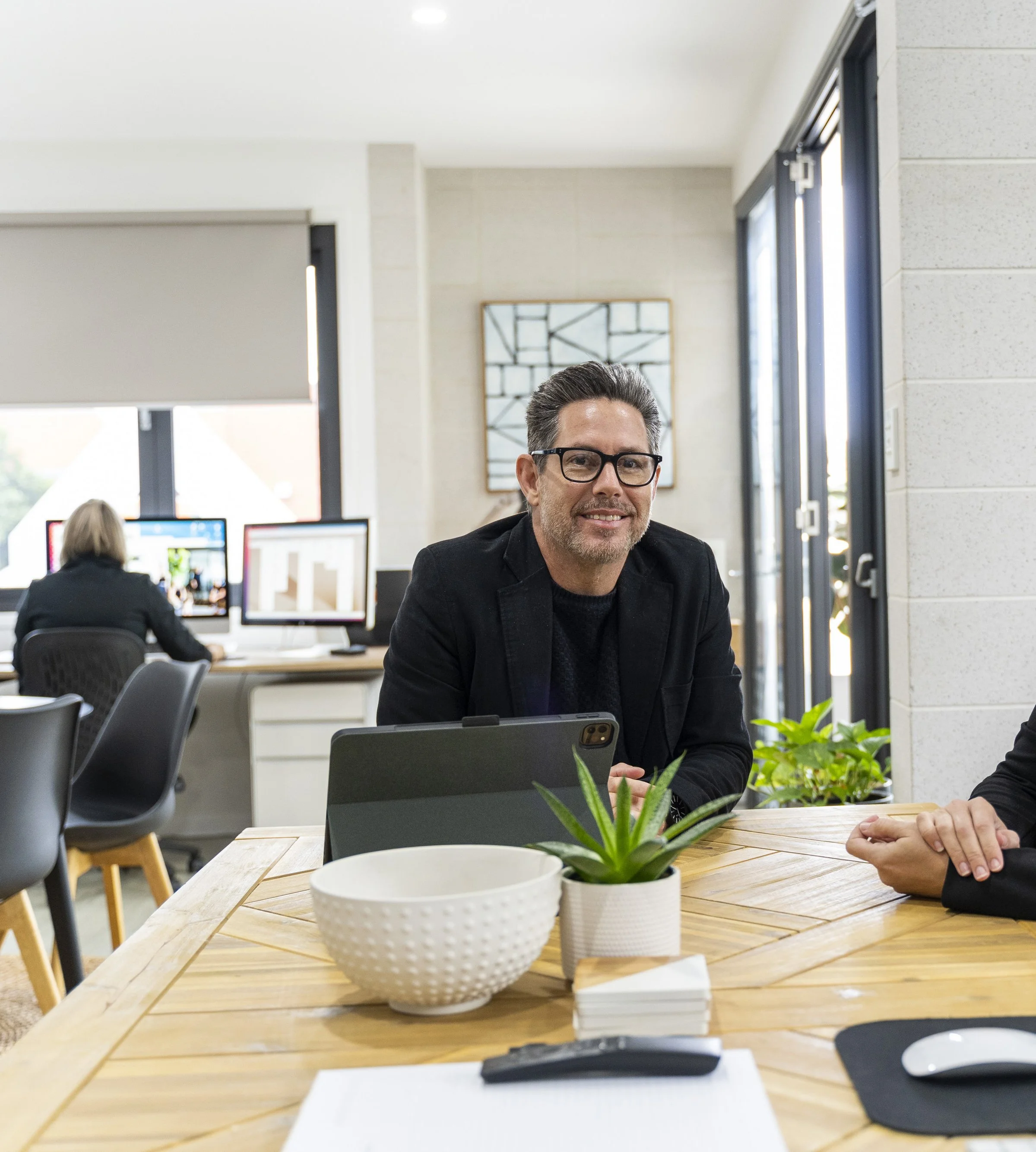 Office interior with desk, potted plant, computer monitors, and people working, including a man wearing glasses smiling at the camera.