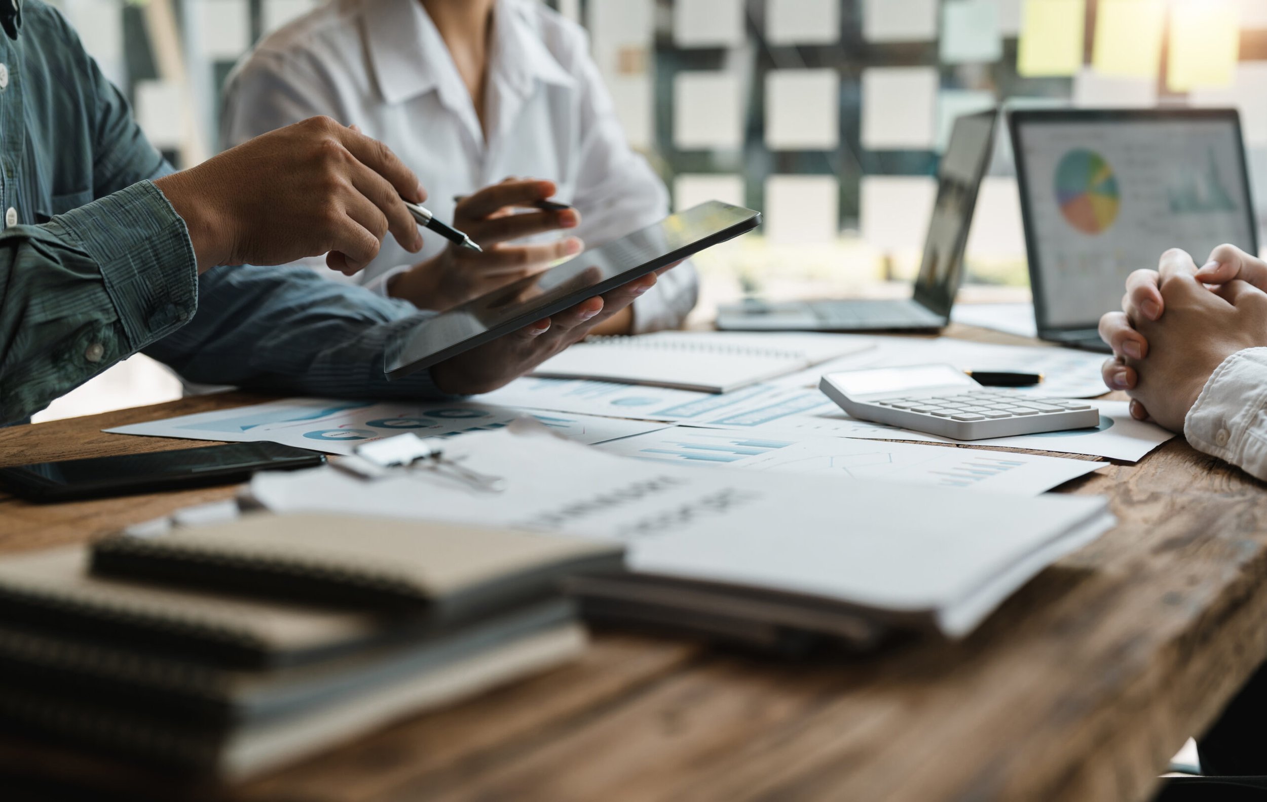 People working together at a wooden table with documents, laptops, and tablets, in a bright office environment.
