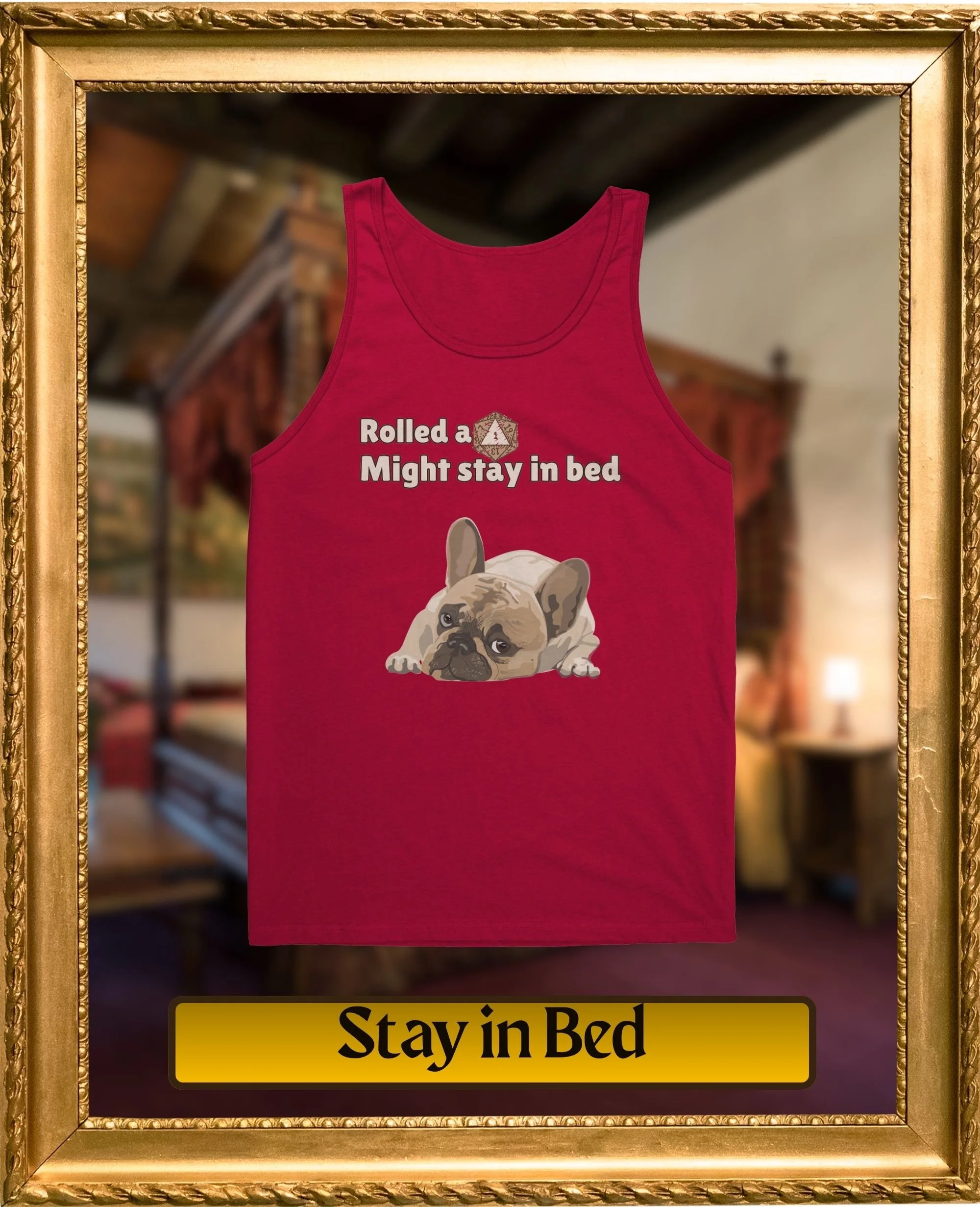 Red tank top with a graphic of a French Bulldog and the text "Rolled a Might stay in bed" on the front, framed by a gold decorative frame. Blurred background of a rustic interior.