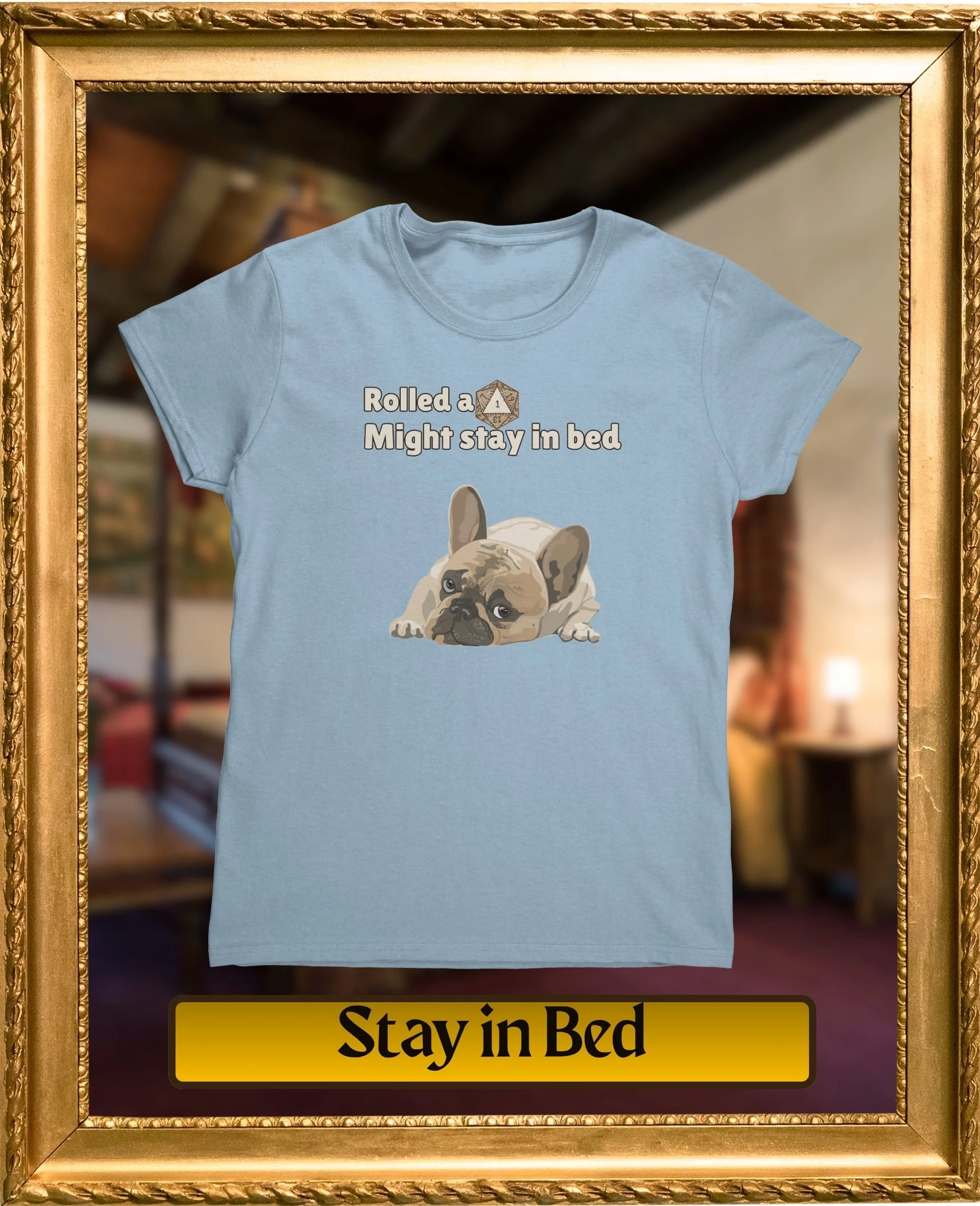 Light blue T-shirt hanging on a display with cartoon illustration of a French Bulldog puppy lying down. The T-shirt has the text "Rolled a 1 Might stay in bed" above the illustration. The background features a blurred store interior.