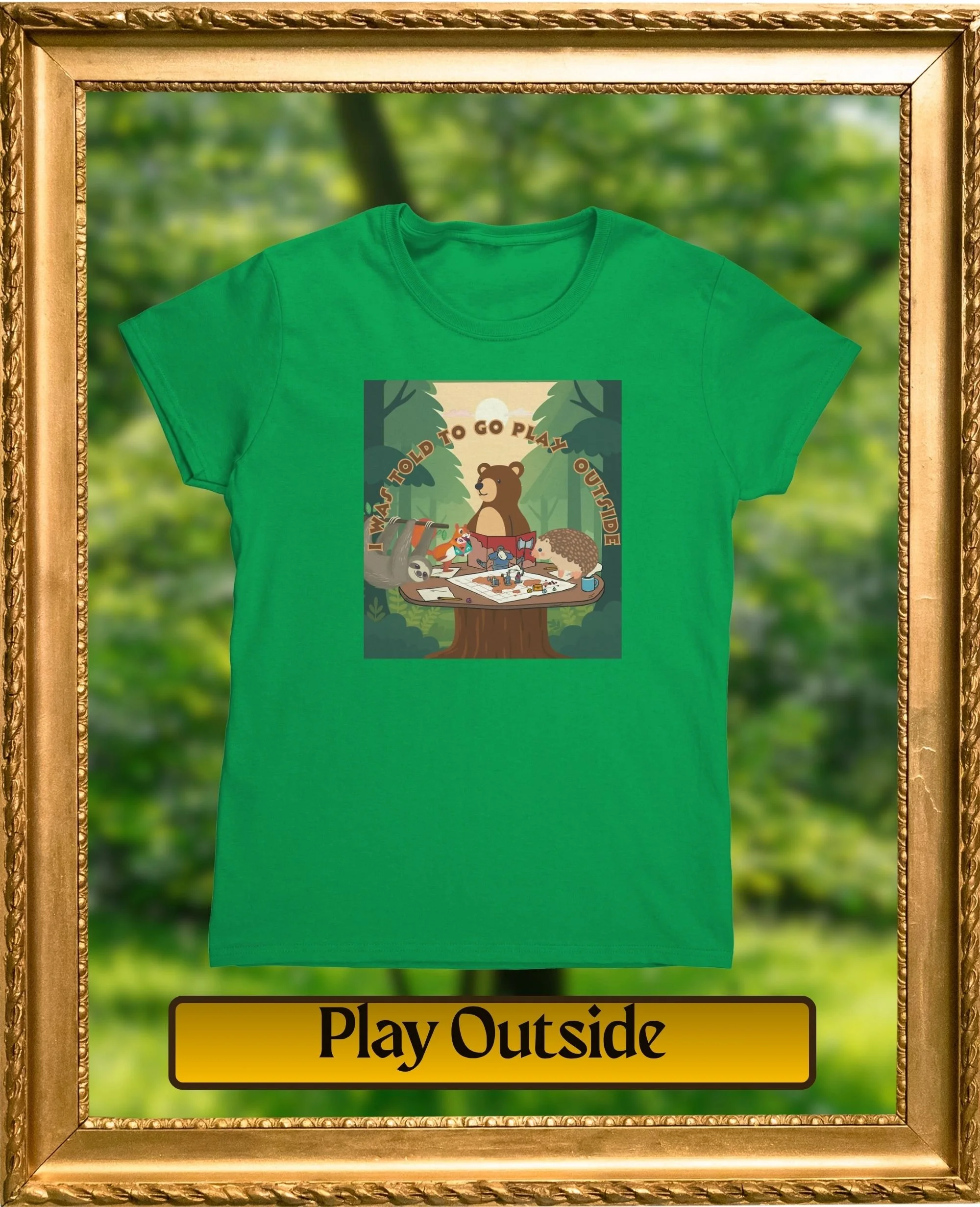 Green T-shirt with a graphic of trees, a bear, a fox, a hedgehog, and camping items, and the text "I WAS TOLD TO GO PLAY OUTSIDE". The T-shirt is displayed inside a decorative gold frame, with a blurred outdoor background.