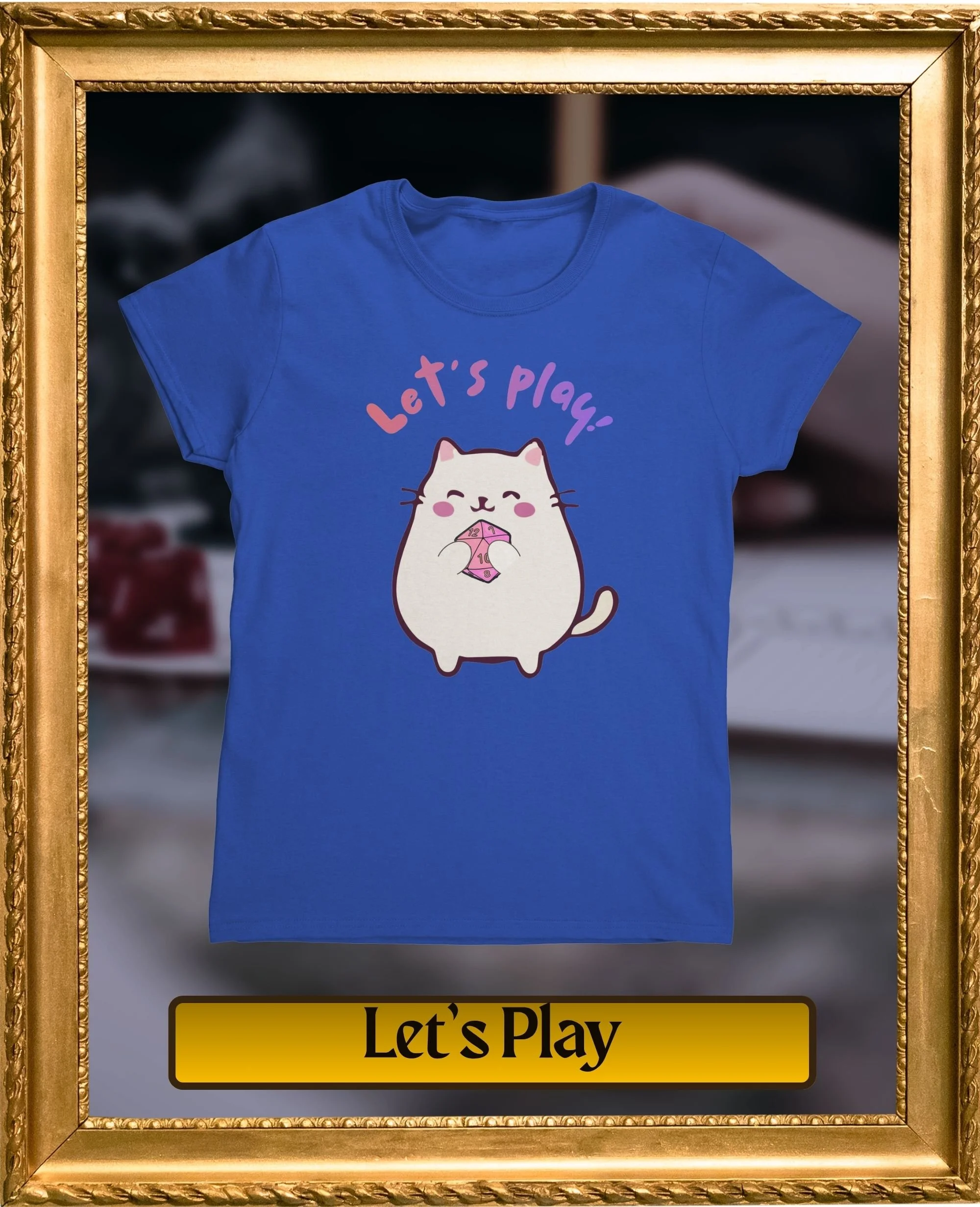 A blue T-shirt with a cartoon hamster holding a pink dice and the text "Let's Play" above it, framed in a gold picture frame, with a blurred background.