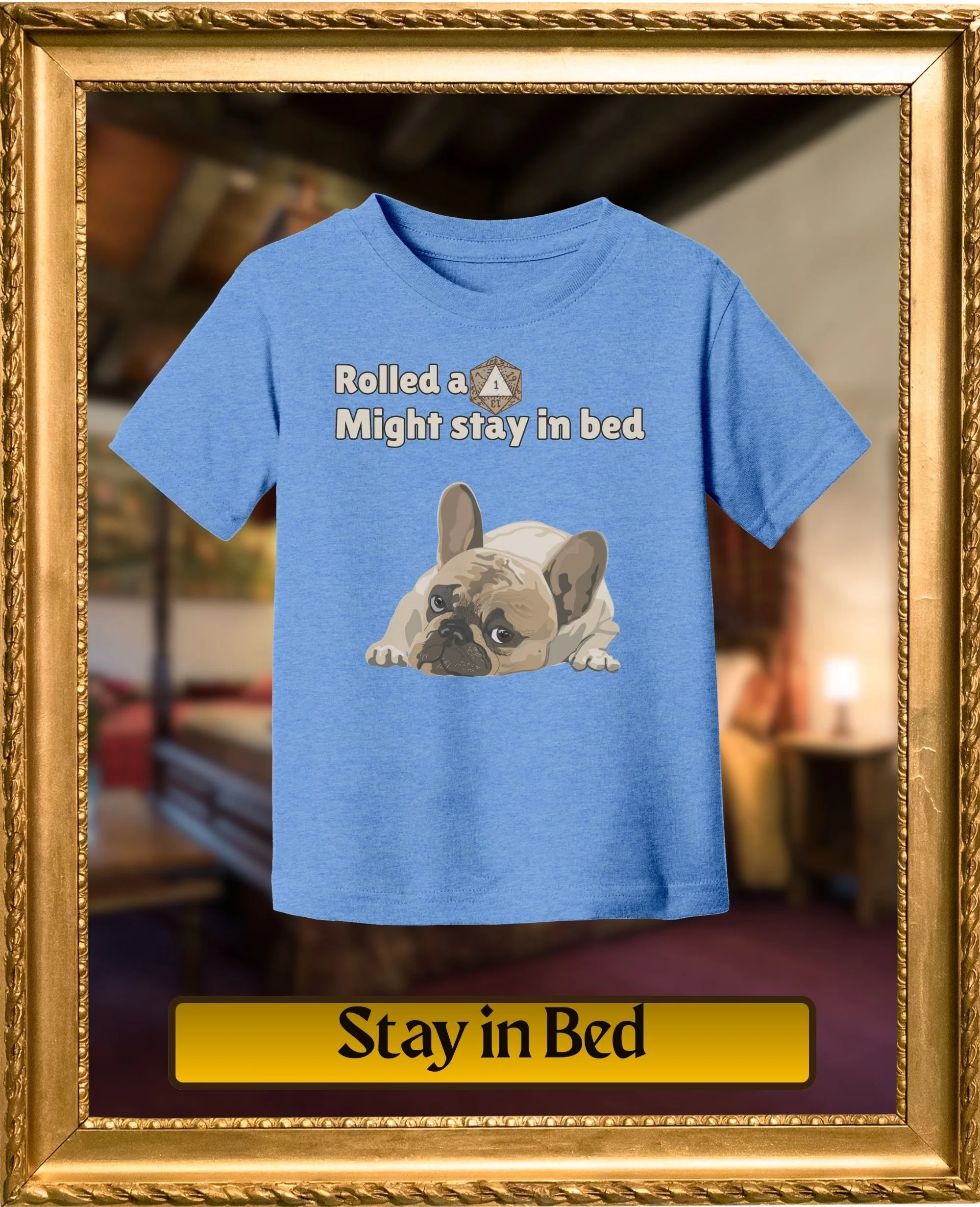 Framed image of a blue t-shirt with a graphic of a French Bulldog and the text "Rolled a Might stay in bed" on it. The background is blurred, and part of a yellow sign with black text "Stay in Bed" is visible at the bottom.