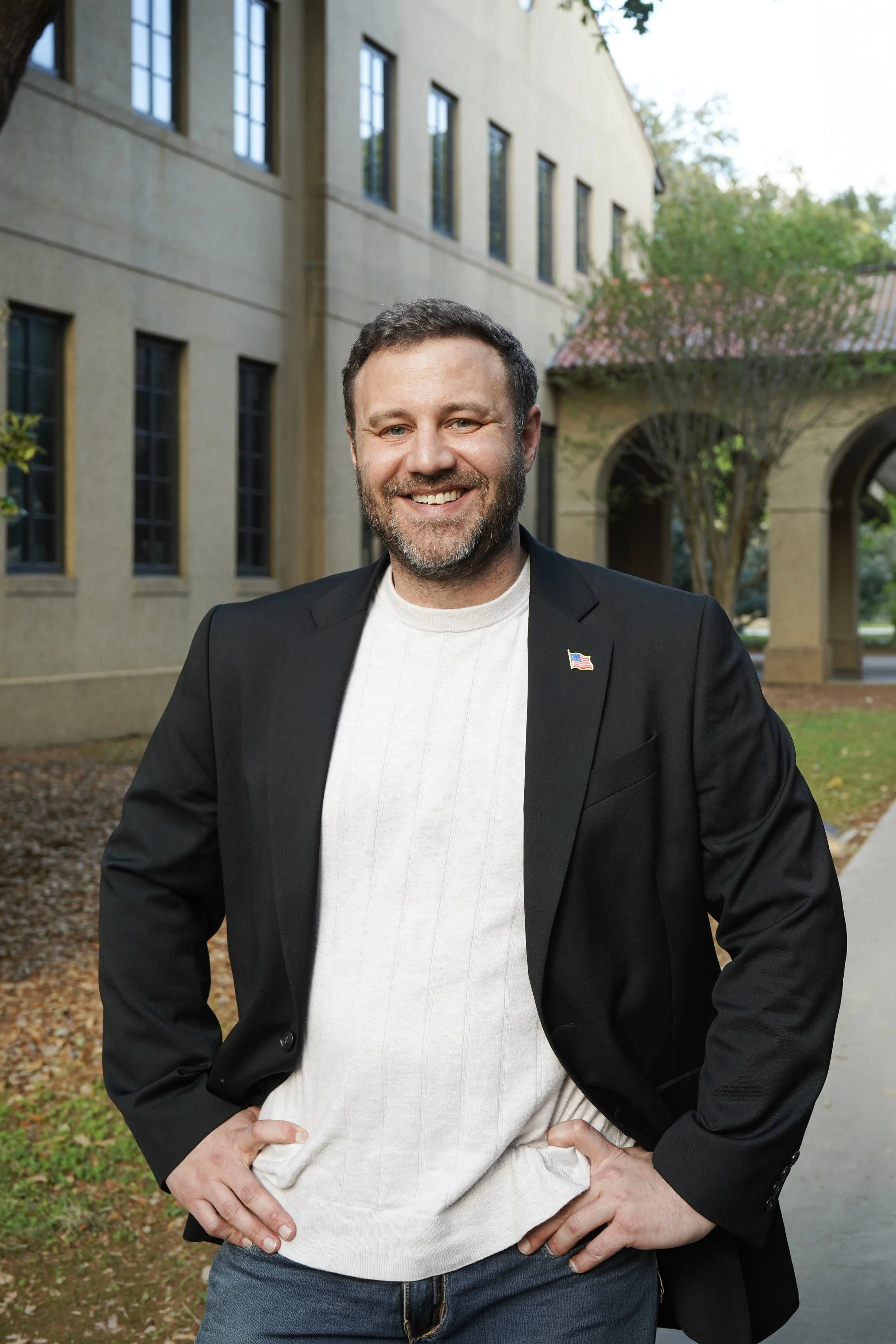 Matt Gromlich, running for congress in Louisiana's 4th district, stands outdoors in front of a building with large windows, wearing a black blazer with a USA flag pin, a white sweater, and jeans, with his hands on his hips.