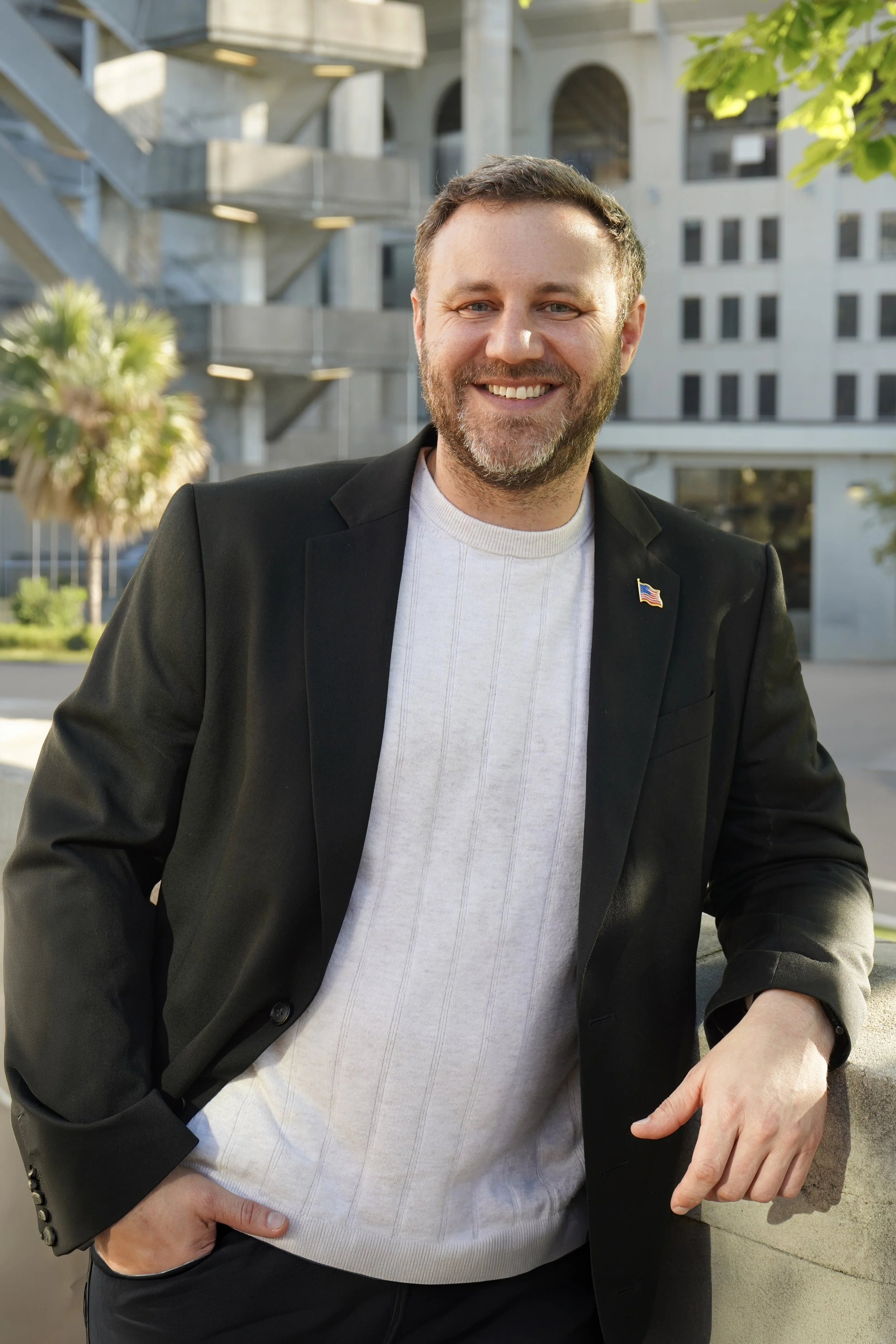 Matt Gromlich, running for congress in Louisiana's 4th district, wearing a black blazer with a small American flag pin, a white textured sweater, and dark pants. He is outdoors with a modern building and trees in the background.