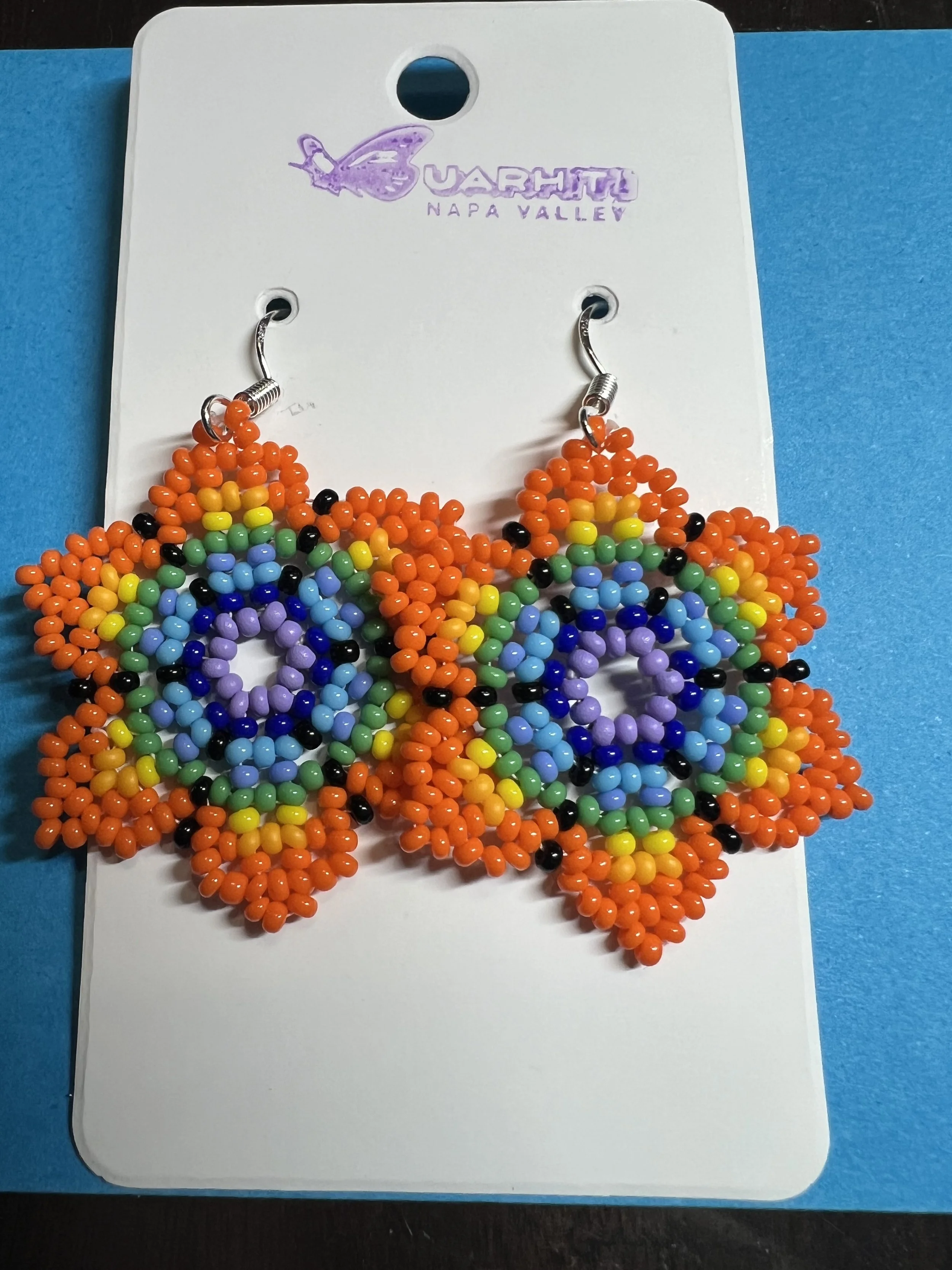 Rainbow-colored beaded earrings displayed on a white card with a purple butterfly logo and text 'Uarthy Napa Valley'. The earrings are made of small beads arranged in rainbow patterns with black beads.