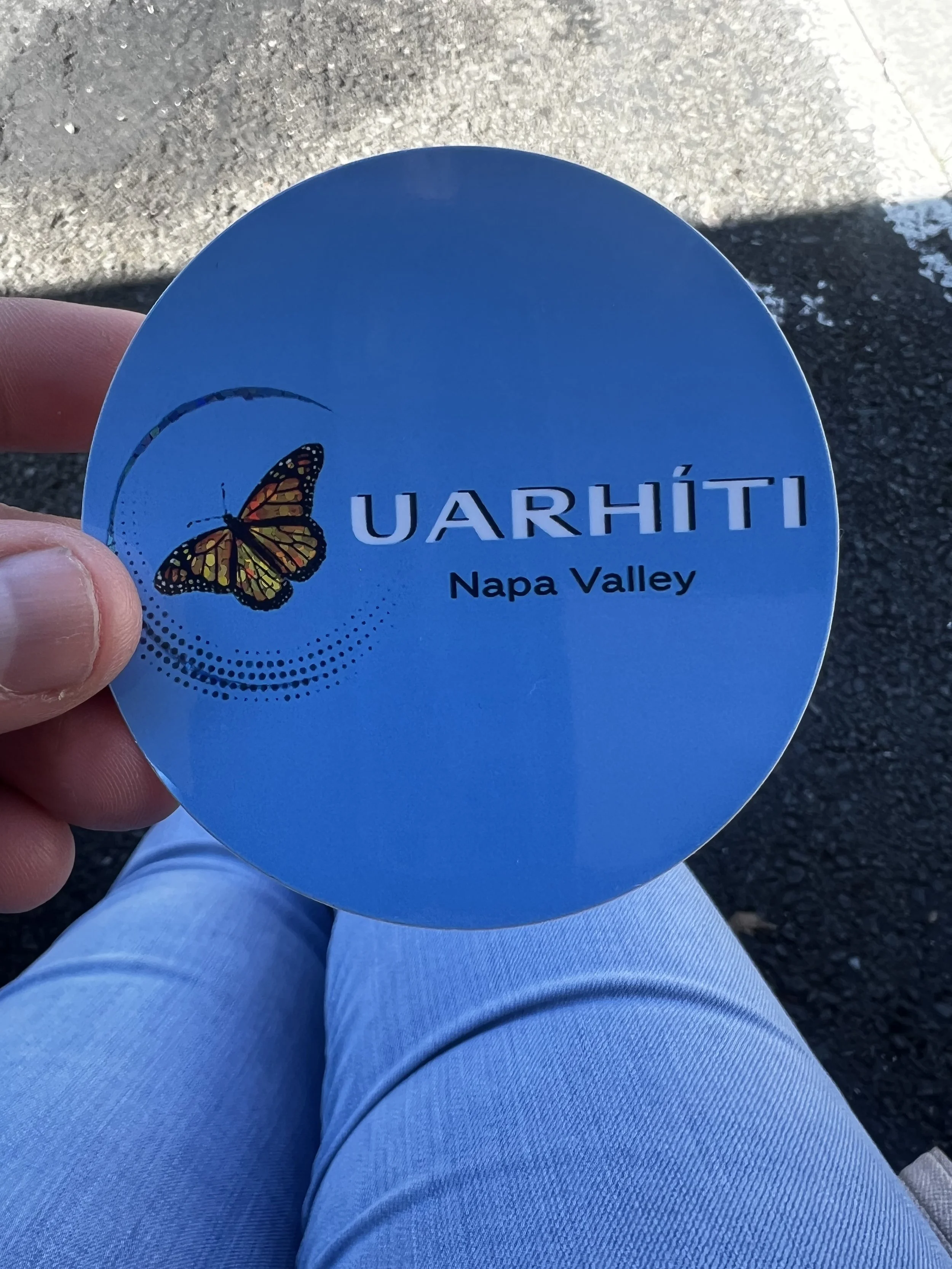 A person holding a blue circular object with a butterfly illustration and the text 'UARHÍT' and 'Napa Valley,' sitting outdoors on a concrete surface.