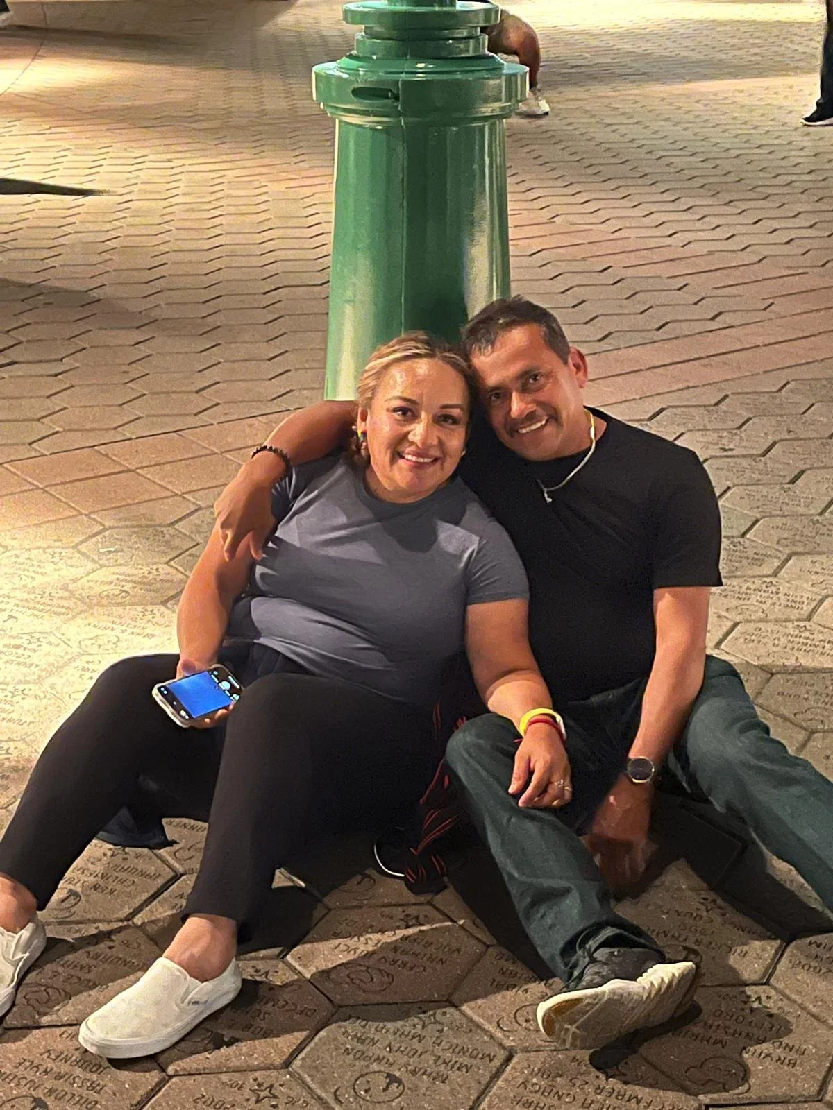 A woman and a man sitting on the ground, smiling, with their arms around each other in front of a green lamp post at night. The woman holds a smartphone, and both are casually dressed.