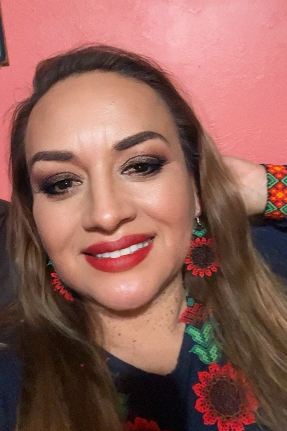 A woman with light brown hair, wearing makeup, smiling, and dressed in traditional embroidered clothing with coral, green, and blue patterns, standing in front of a pink wall.