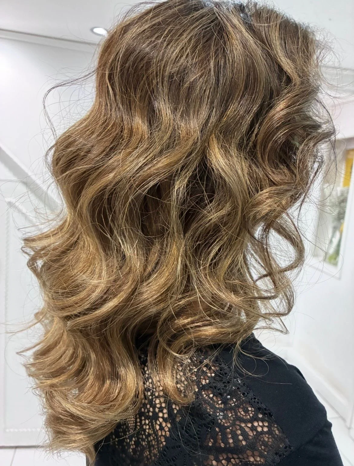 Wavy, light brown hair with highlights styled in loose curls, worn by a person in a black lace top.