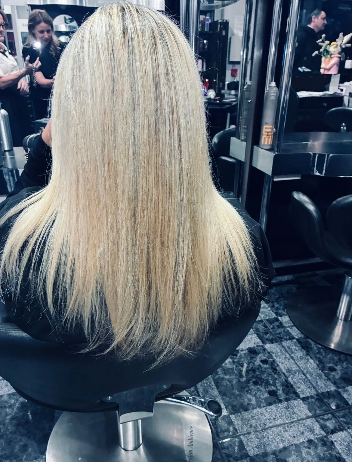 Blonde hair sitting in a salon chair in front of a mirror, with other people and salon equipment visible in the background.