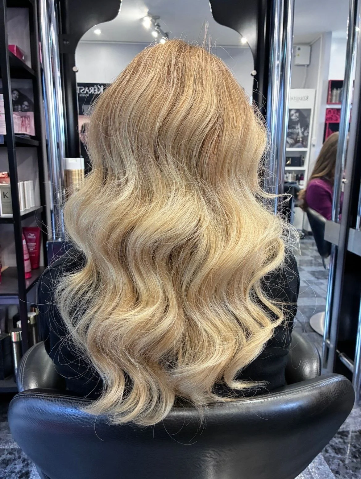 A woman with long, wavy blonde hair sitting in a salon chair.