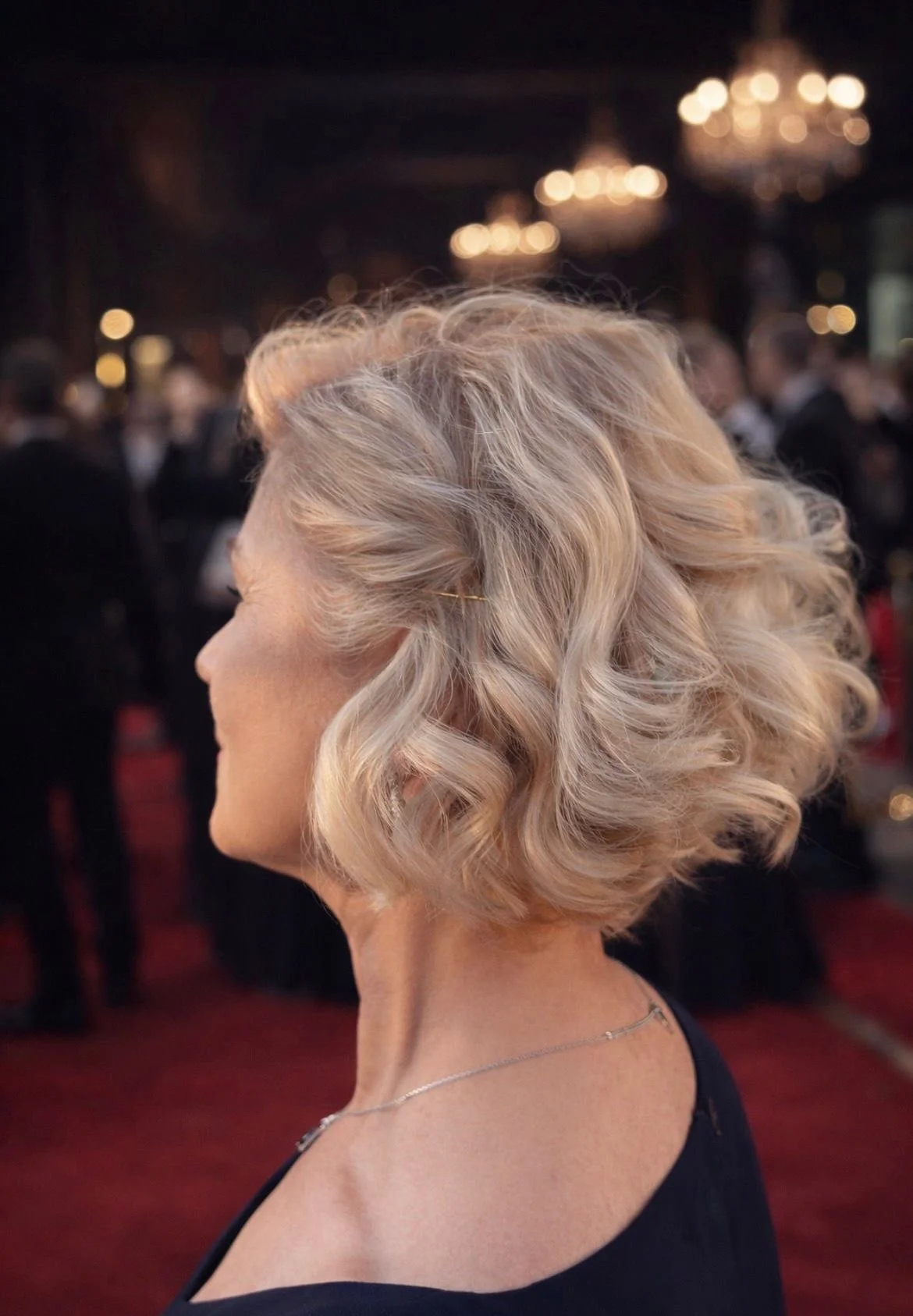 Side profile of a blonde woman with short, curly hair at an indoor event with chandeliers and people in the background.