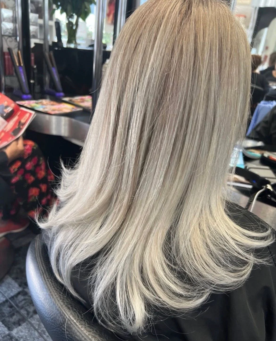 A person with long, straight, platinum blonde hair sitting in a salon chair. The hair has a smooth, shiny texture with soft waves at the ends, and the background shows salon mirrors, chairs, and a person reading a magazine.