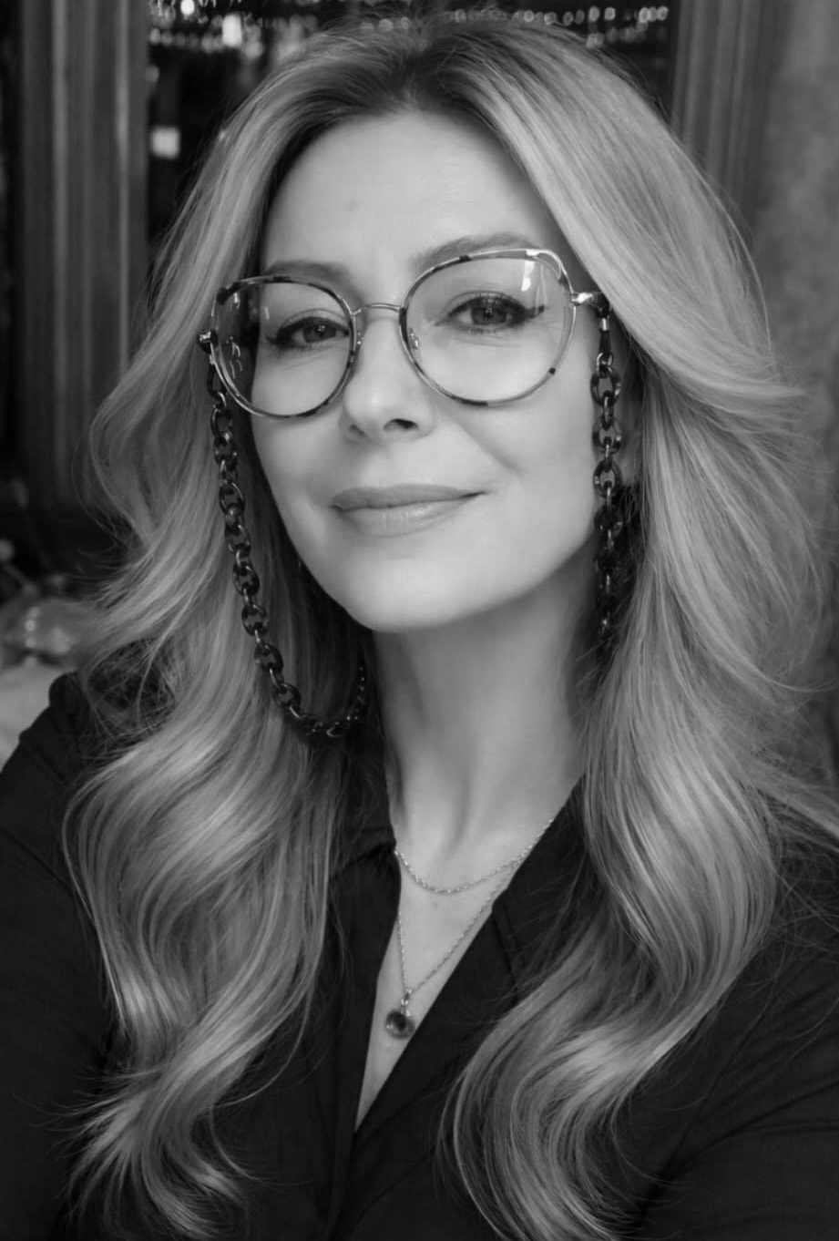A woman with long, wavy hair, wearing glasses with chains, smiling, and dressed in a dark blouse, in a black-and-white photo.