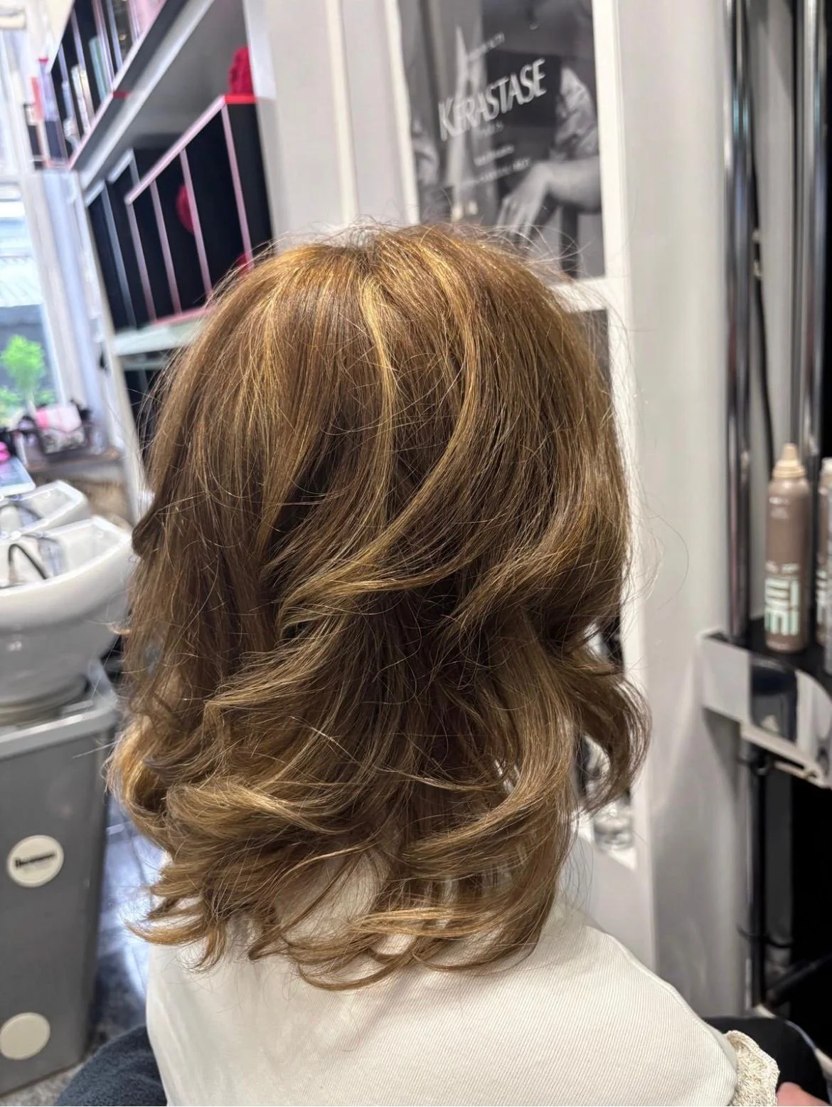 Back of woman with light brown, shoulder-length curly hair inside a hair salon.