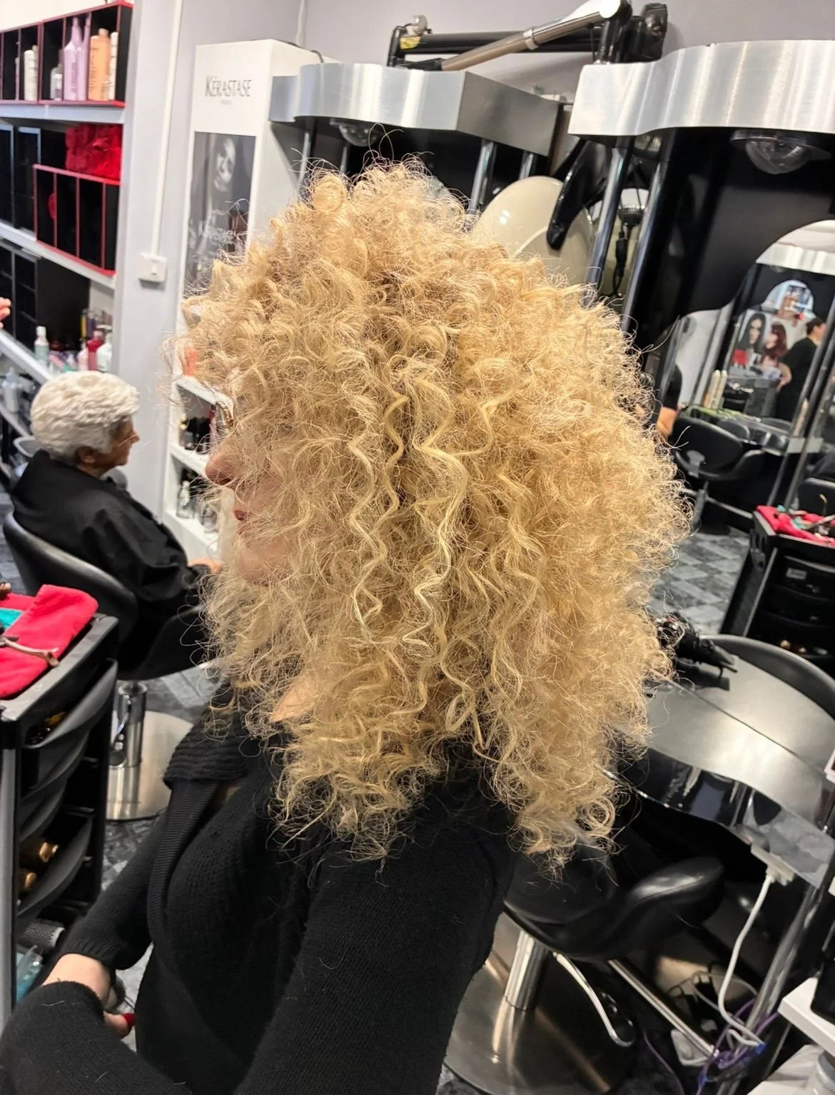 A woman with tight blonde, curly hair sitting in a salon chair.