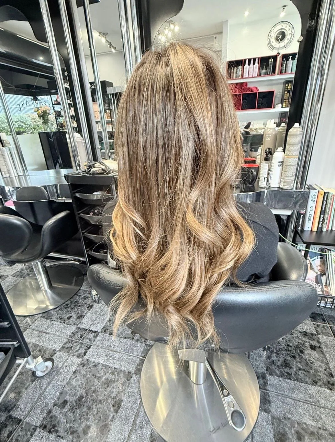 Back view of a woman with long, wavy, blonde hair sitting in a salon chair.
