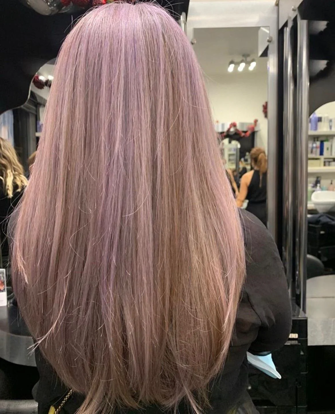 Back view of a woman in a salon with pastel purple and light pink hair, sitting in front of a mirror, with salon equipment and other people in the background.