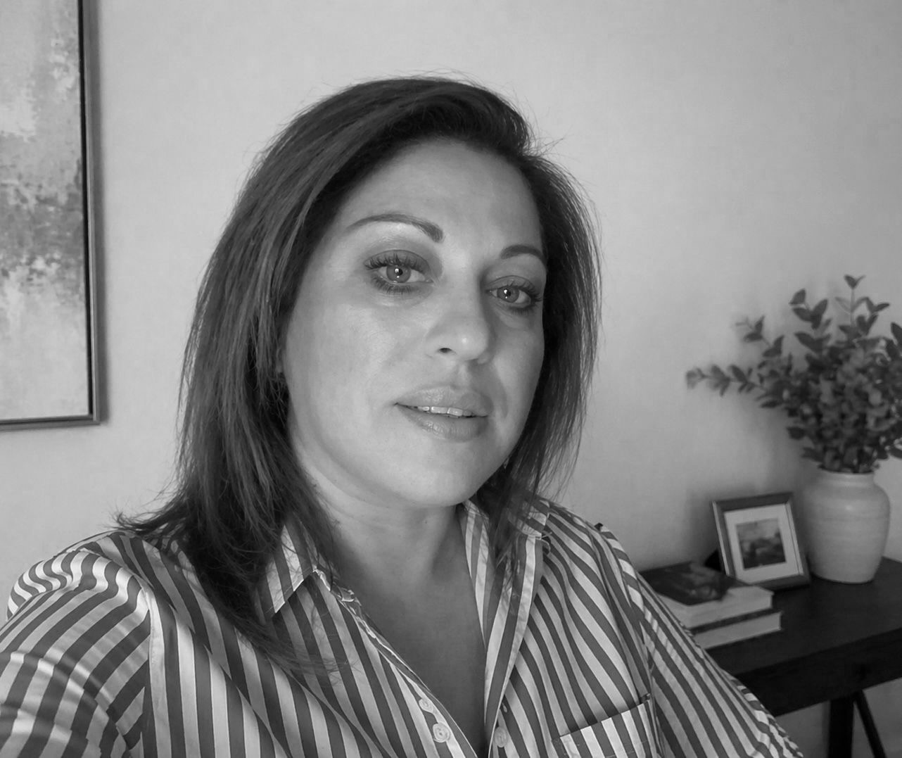 Black and white portrait of a woman with shoulder-length hair, wearing a striped button-up shirt, sitting in a room with a table, a vase with a plant, and framed photographs in the background.