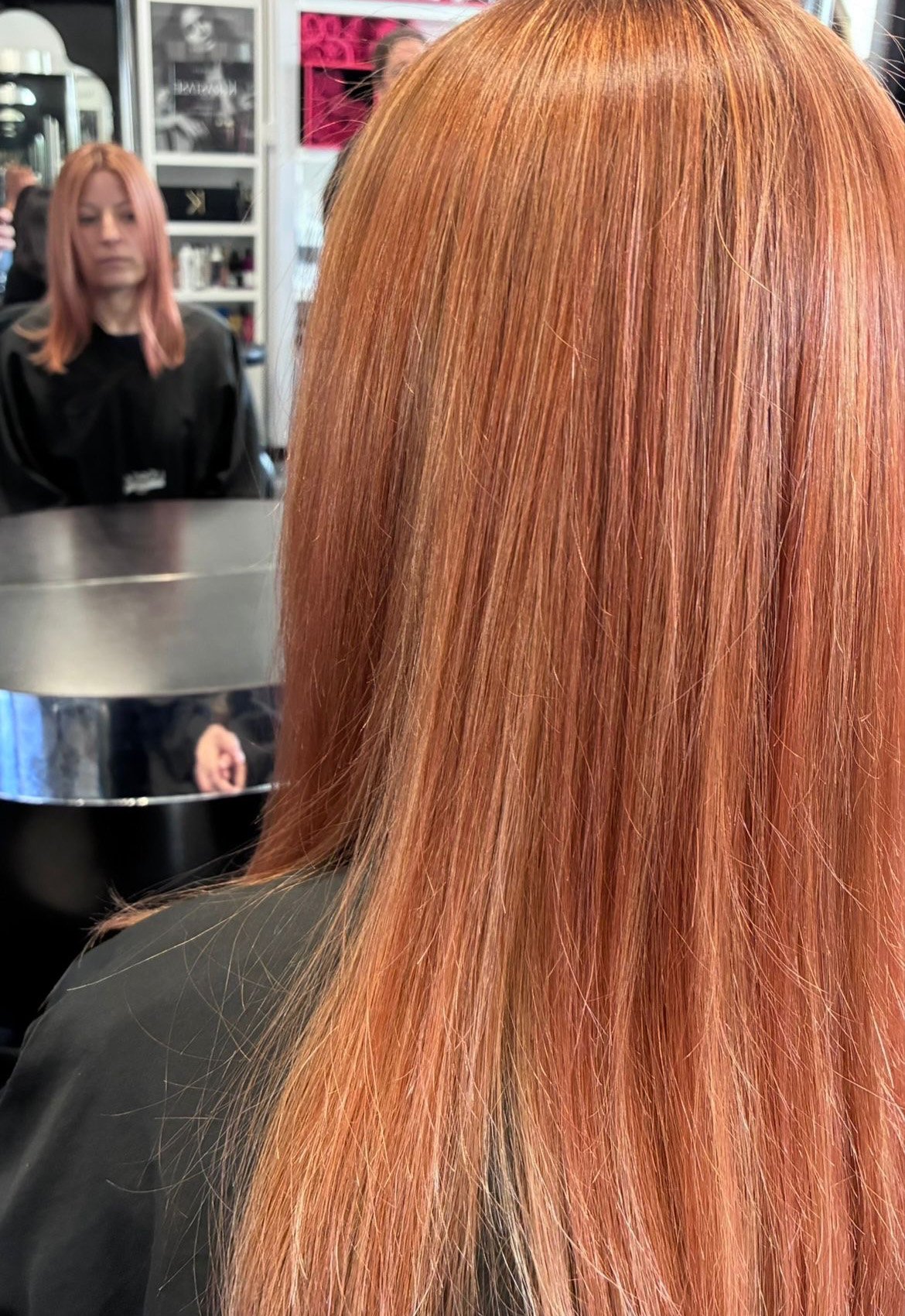 Woman with long, straight, orange-red hair in a salon mirror reflection, wearing a black cape, with salon products and photos in the background.