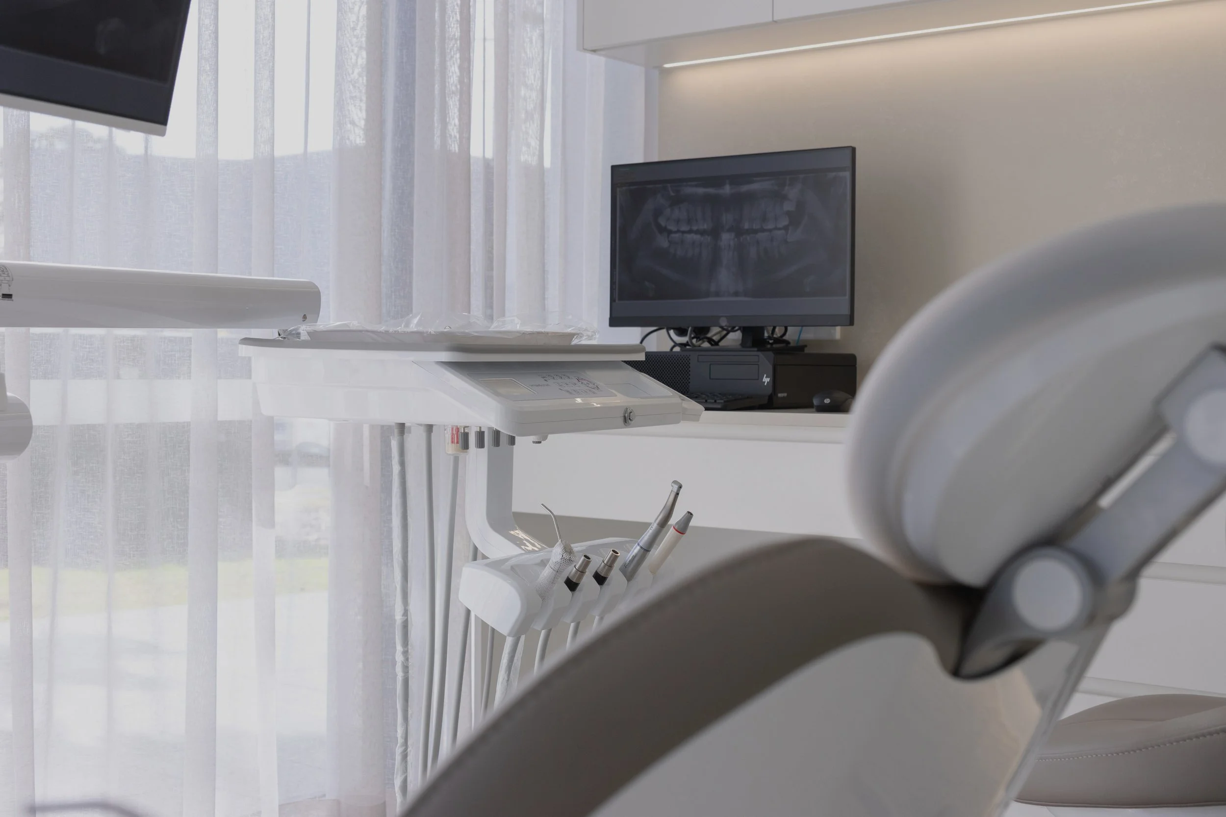 A dental examination room with a dental chair, dental tools, a computer monitor displaying a dental X-ray, and a window with sheer curtains.