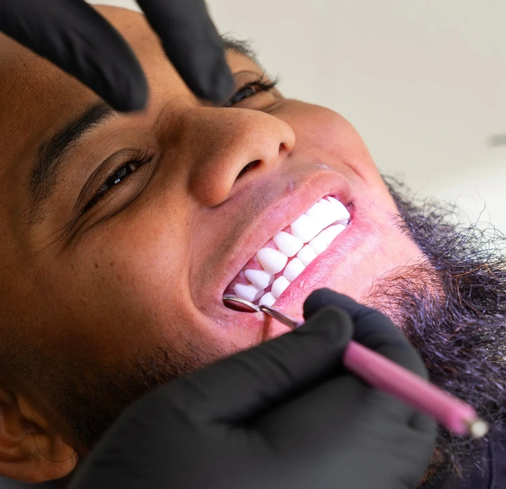 A man lying in a dental chair, smiling, as a dentist examines his teeth with dental tools.
