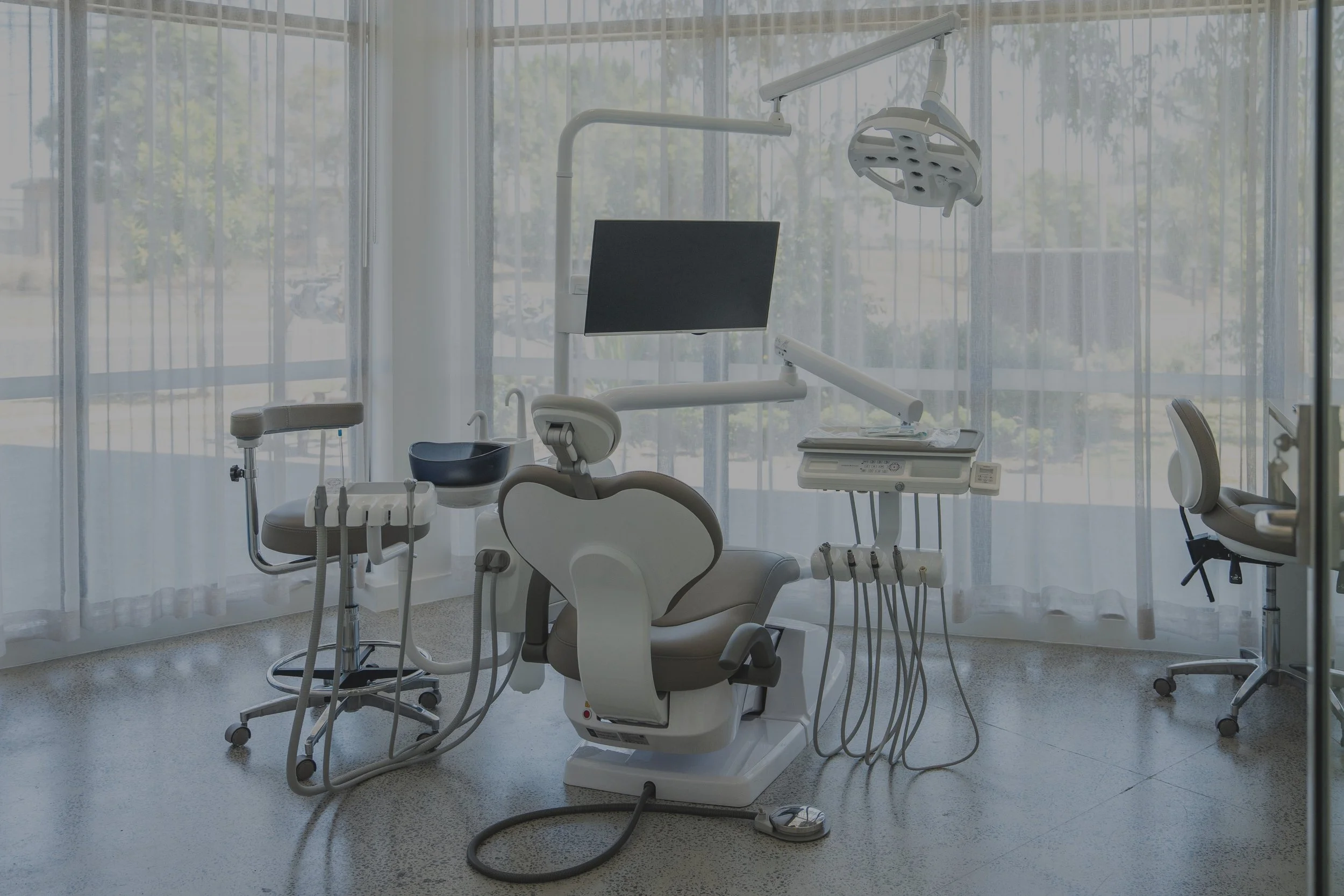 A modern dental office with a dental chair, monitor, and various dental equipment, all in a room with large windows and sheer curtains.