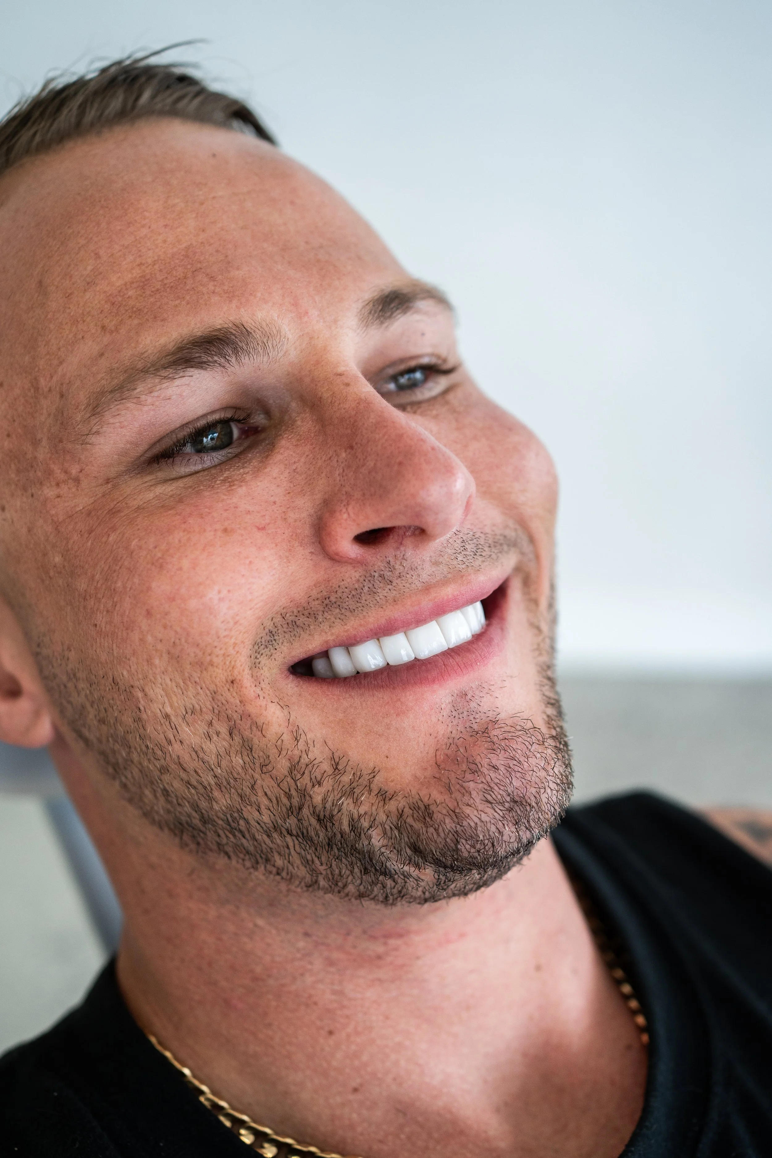 Close-up of a smiling white man with short hair, a beard, and blue eyes, wearing a black shirt and a thin gold chain, against a light background.