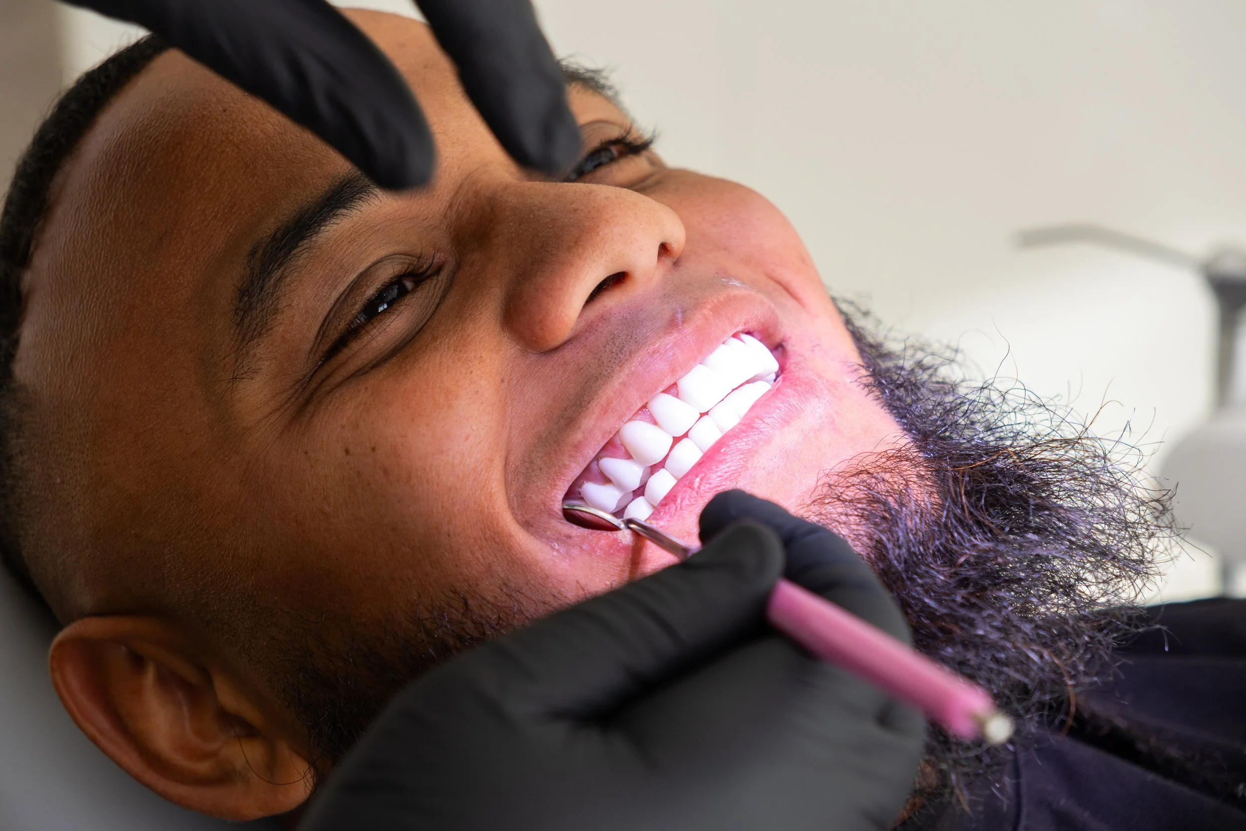 A man lying in a dental chair, getting his teeth examined by a dentist wearing black gloves, using dental tools.