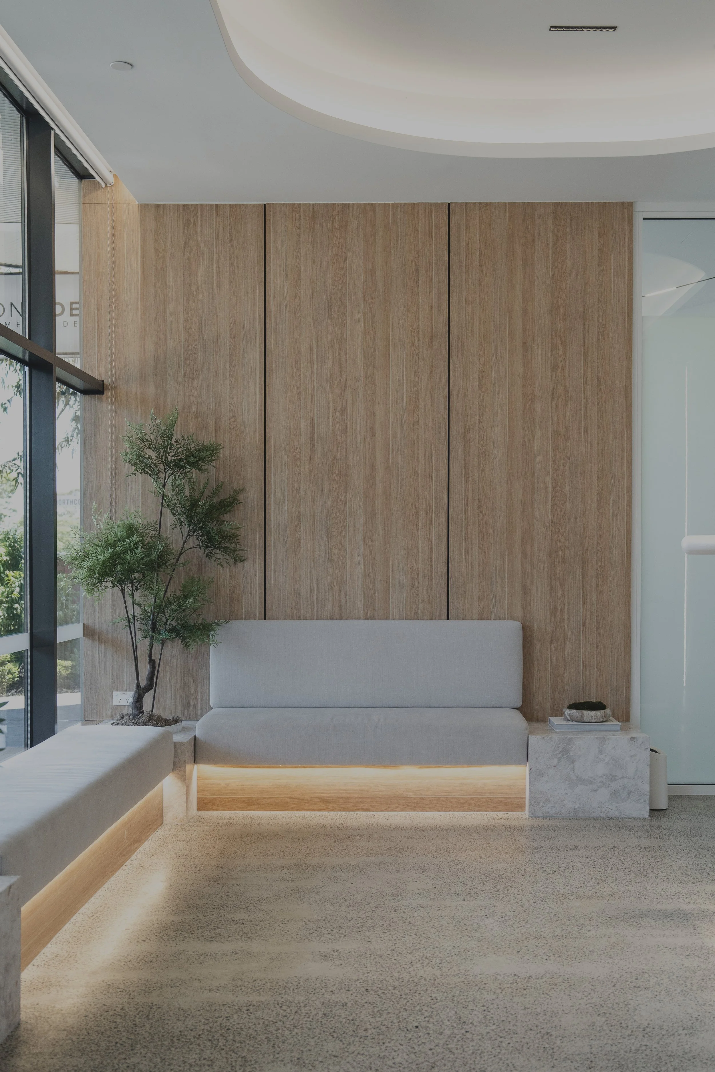 Modern lobby with a light grey couch, a potted plant, and a wooden wall panel. The space features minimalist decor and ambient lighting.