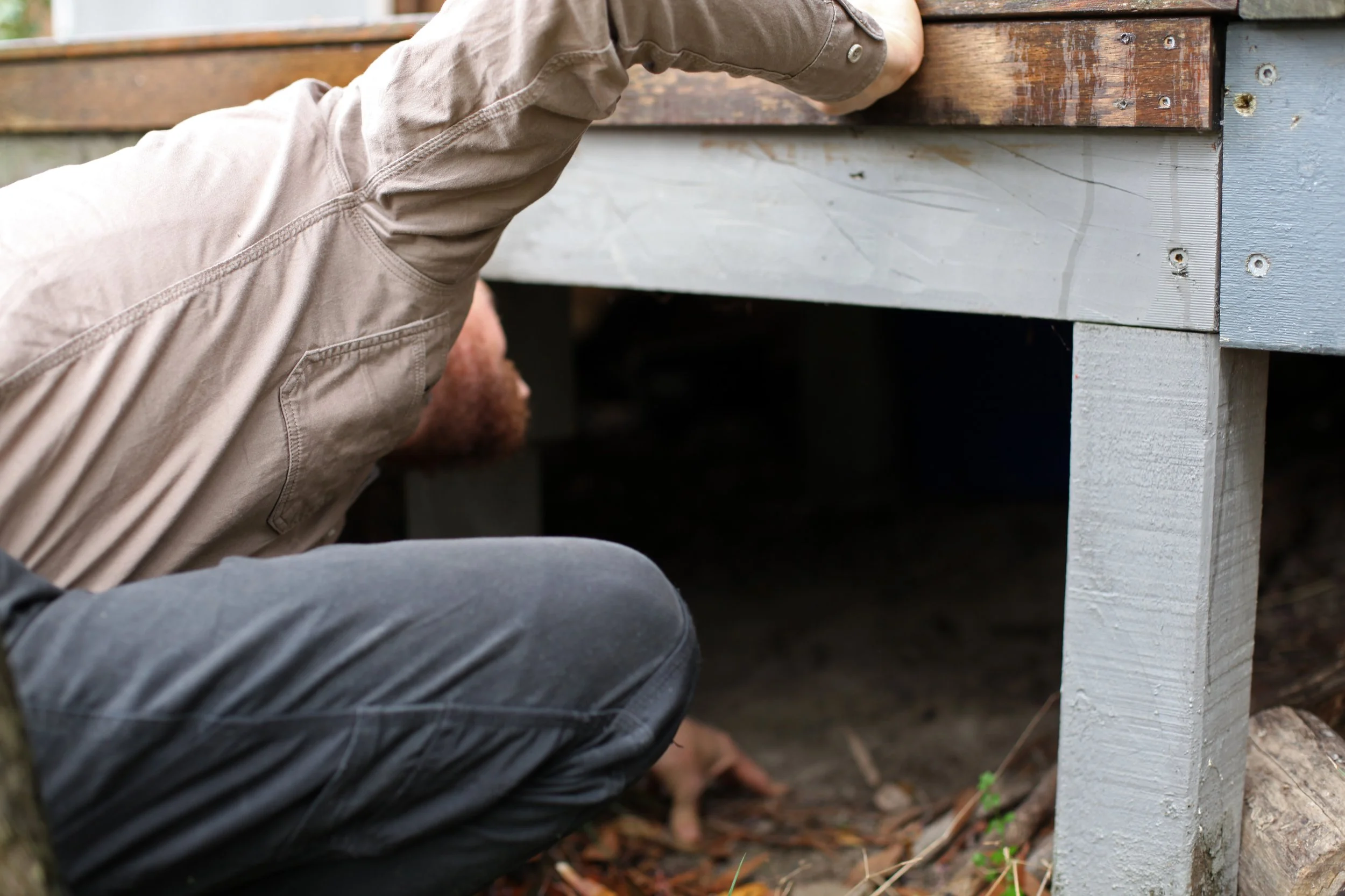 A man wearing a beige shirt and grey pants crouching under a wooden deck, inspecting the structure. The deck is supported by wooden posts, with the ground covered in dirt and some green plants.