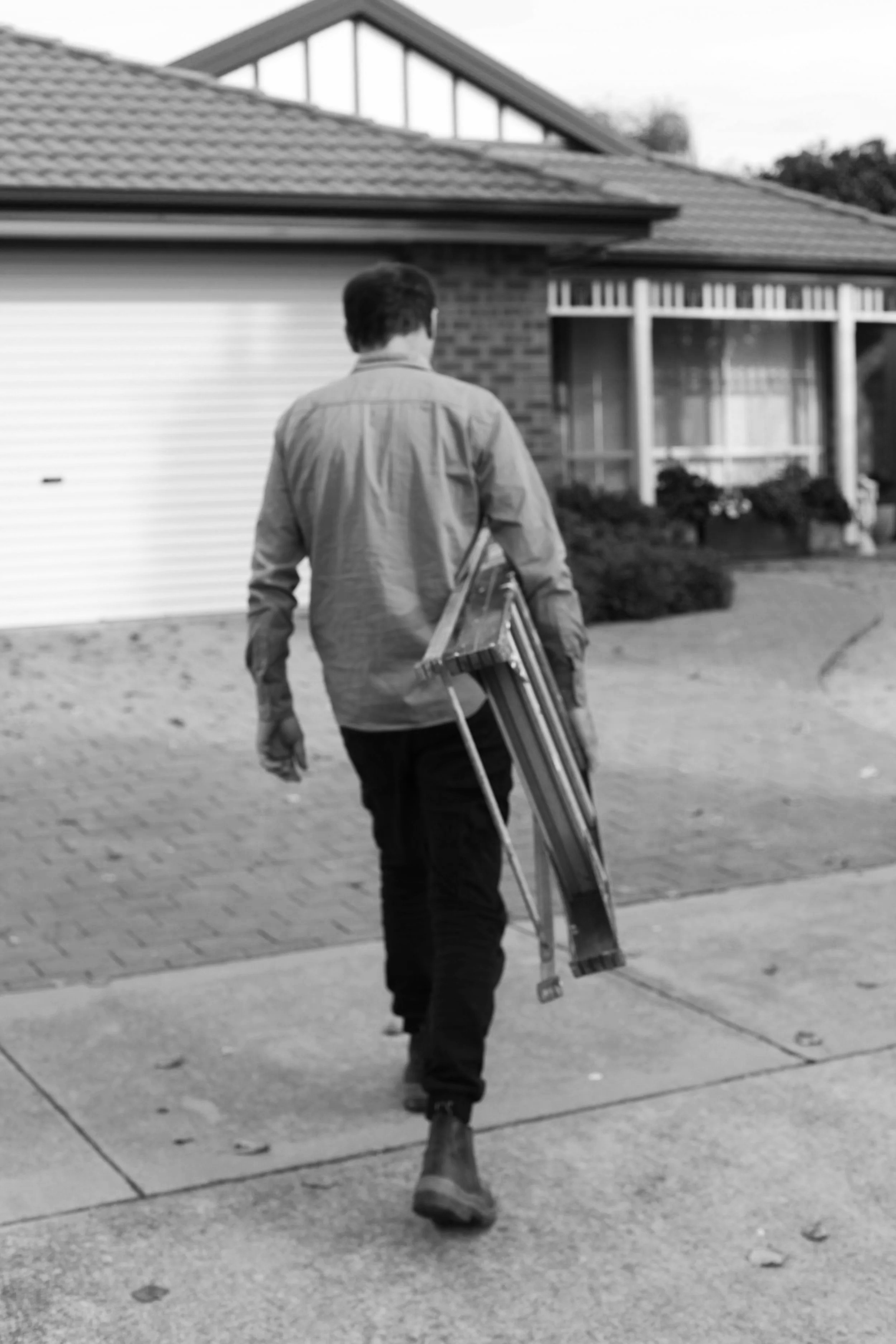 A building inspector walking towards a house carrying a ladder, facing away from the camera, with a house and garden in the background.
