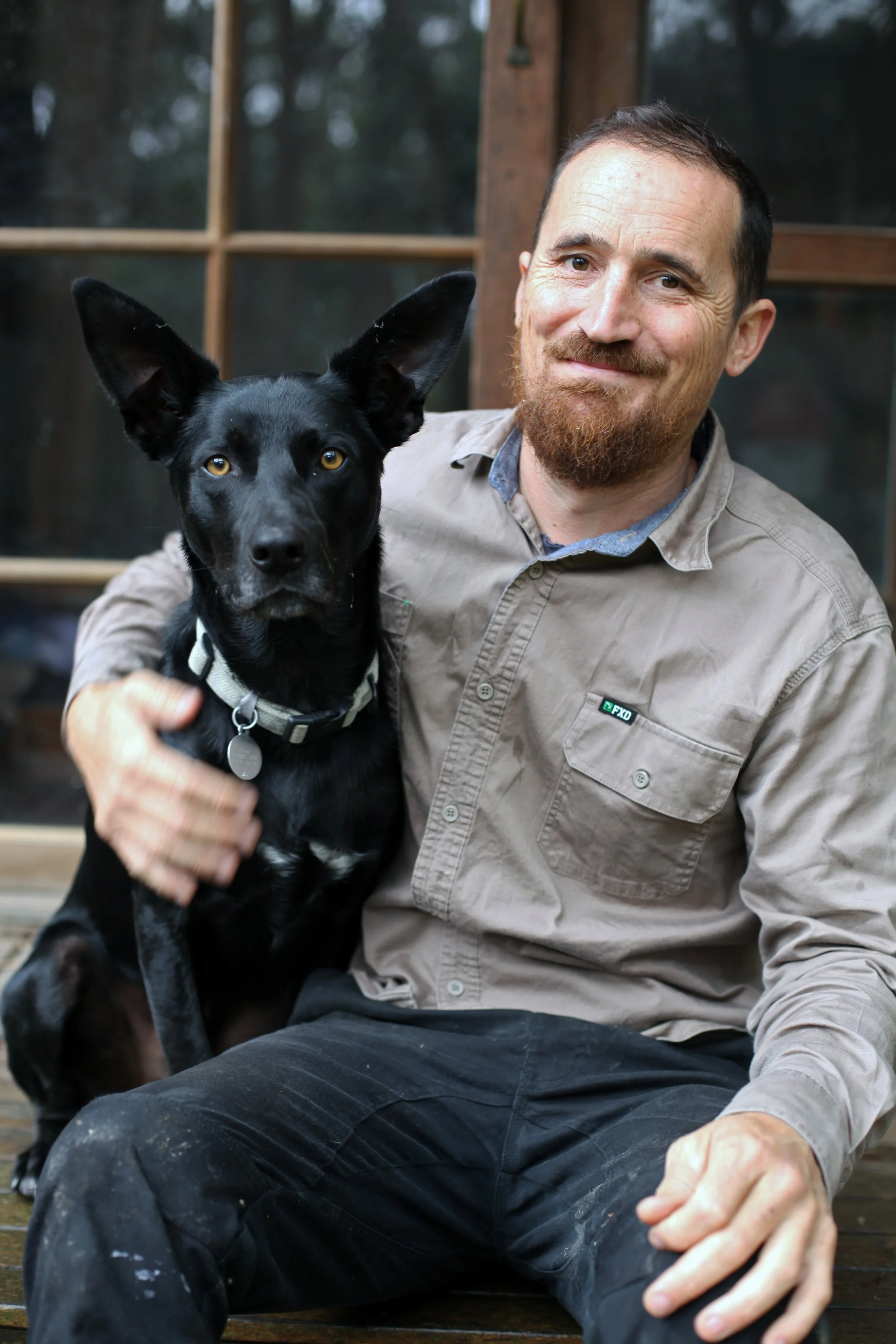 A man with a beard smiling, sitting on a wooden deck with a black kelpie with brown eyes.