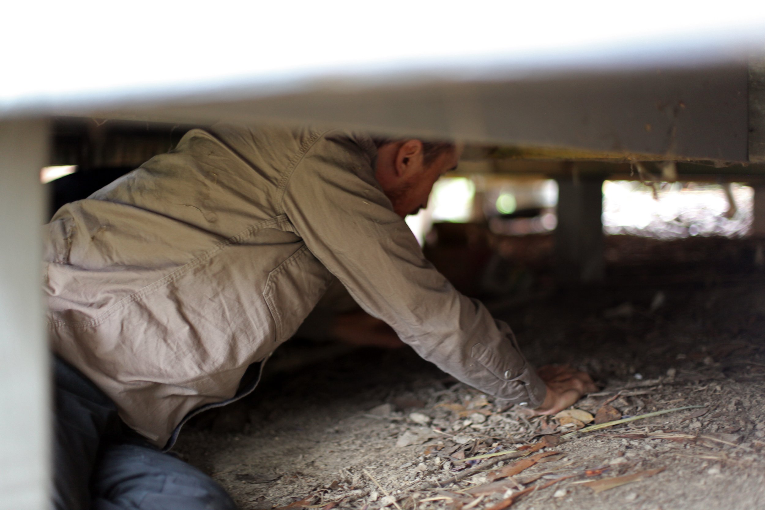 A man crawling under the sub-floor of a house, with dirt and leaves on the ground.