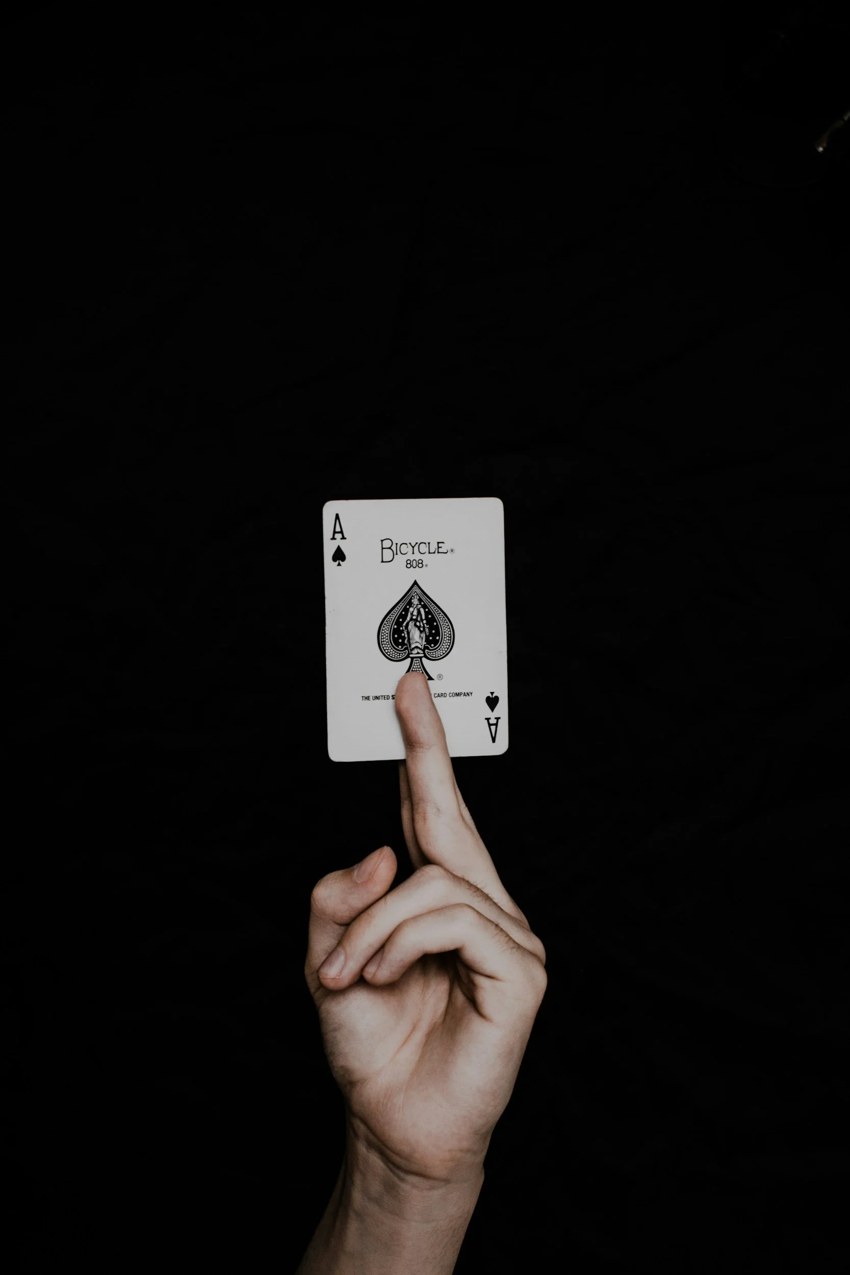 A person holding an Ace of Spades playing card upright with their index finger, against a black background.