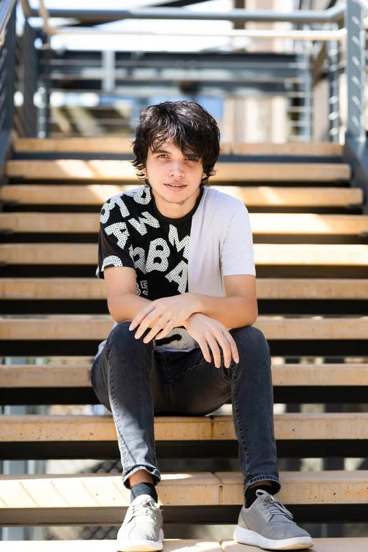 A young man with dark, curly hair sitting on wooden stairs outdoors, looking at the camera with a relaxed expression, wearing a black and white graphic T-shirt, black jeans, and gray sneakers.