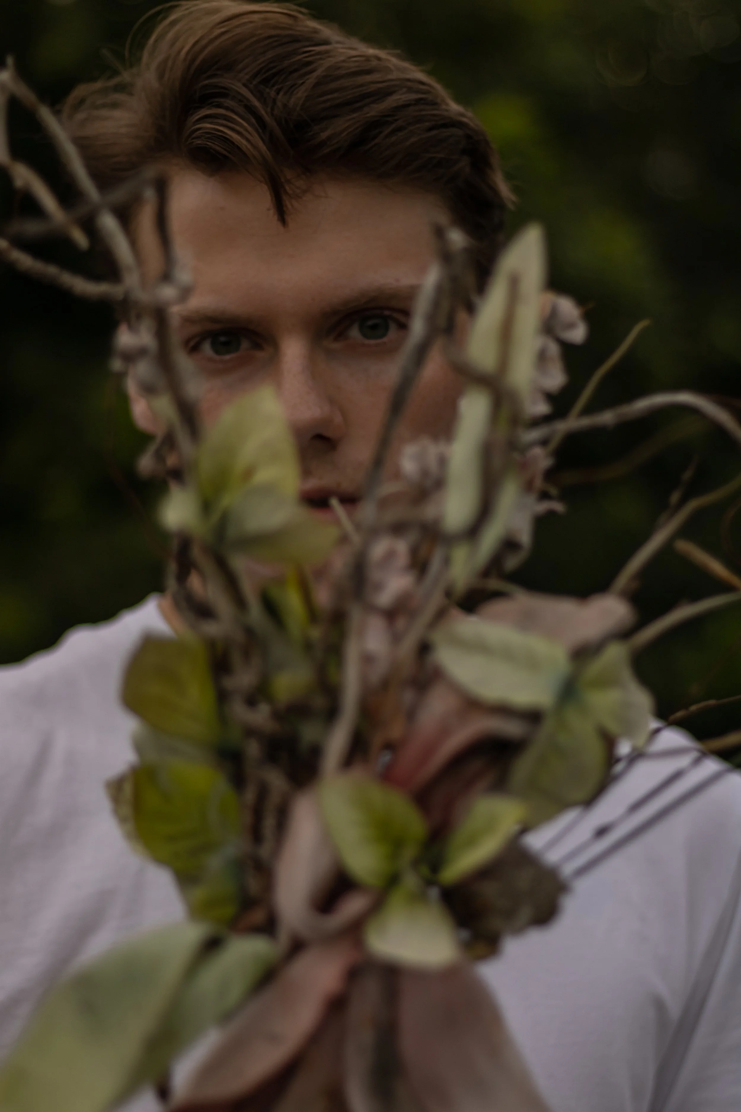 A person with light brown hair and wearing a white shirt holding a plant with green leaves and branches in front of their face, partially obscuring it.