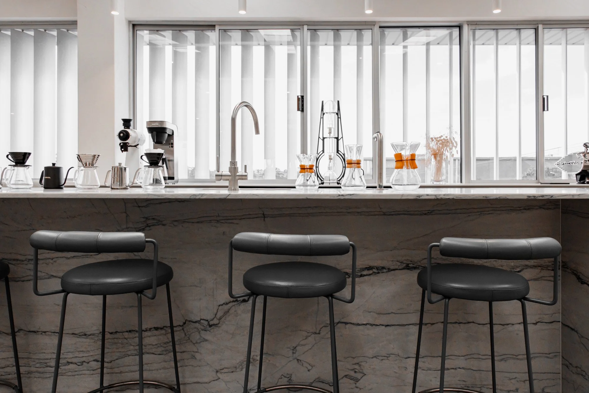 Modern cafe kitchen with marble counter, three black bar stools, large window with vertical blinds, coffee brewing equipment, glass Chemex coffee makers, and decorative items.