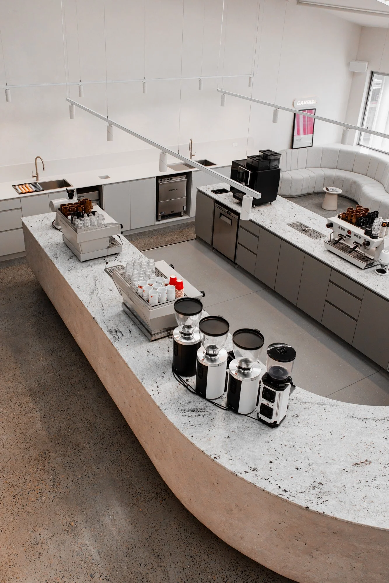 Modern coffee shop or cafe with coffee brewing equipment, grinders, and cups on a curved marble countertop, seating area with white curved banquette along window, and minimalist decor.