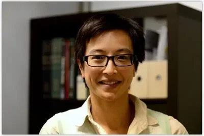 A woman with short dark hair, glasses, and a light-colored shirt smiling in front of a bookshelf.