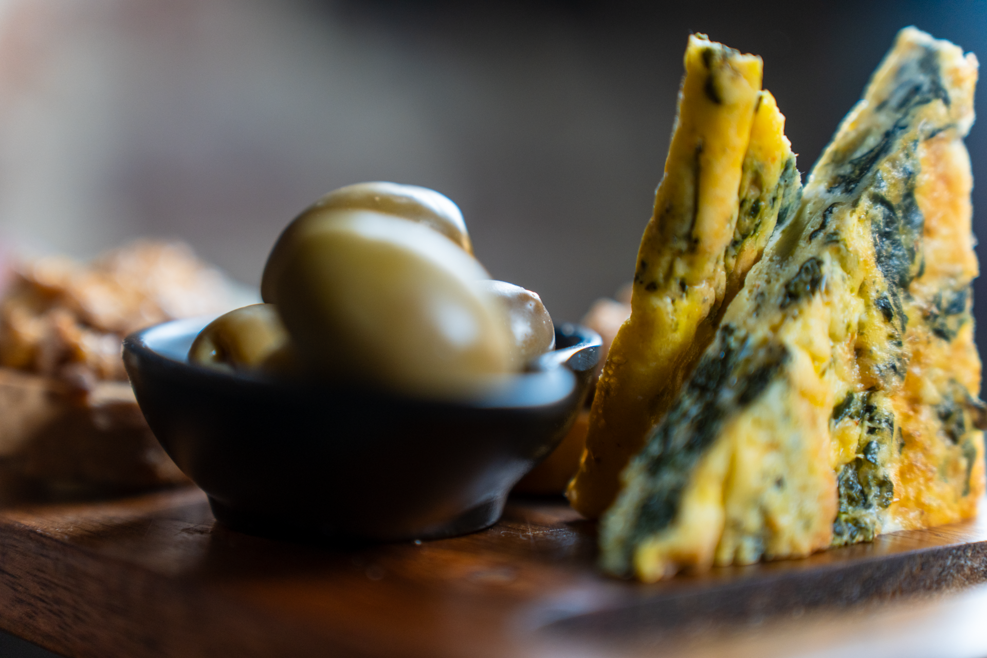 Fette di frittata con spinaci e una ciotola di olive su un tagliere di legno.