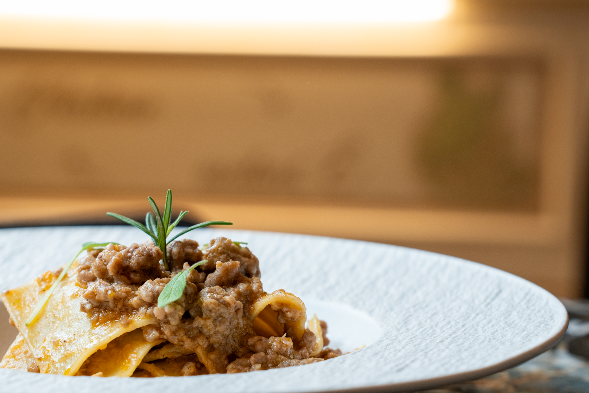 Pasta fatta a mano con ragù di carne, decorata con rosmarino e foglie di salvia, servita in un piatto bianco.