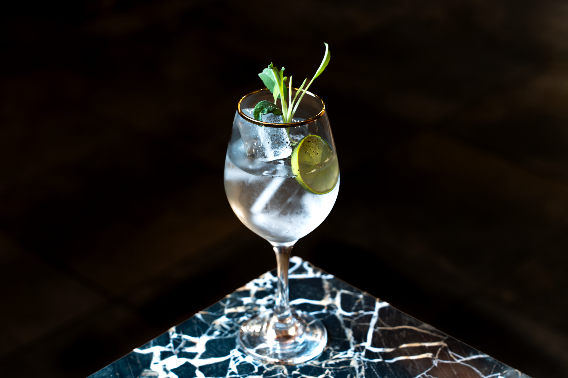 Un bicchiere di cocktail trasparente con Gin, lime, ghiaccio e foglie di menta, posizionato su una superficie in marmo nero con venature bianche.