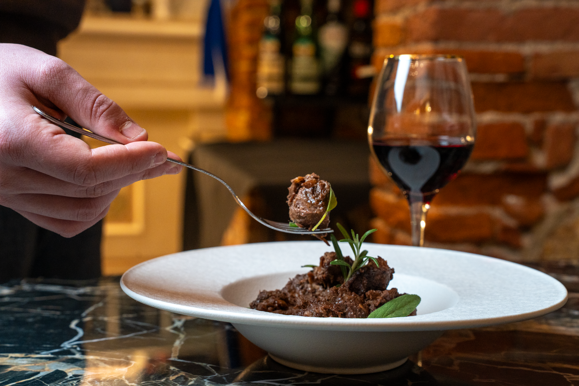 Una mano conforando un piatto con cibo, una forchetta con un boccone, un bicchiere di vino rosso e un murale di mattoni dietro.